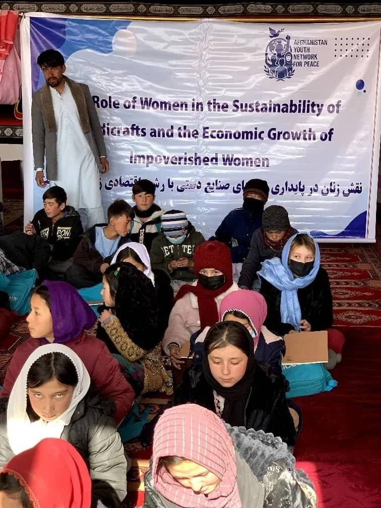 Group of children and young adults sitting on the floor in front of a banner, attending a workshop or seminar about women's role in sustainability and economic growth, organized by Afghanistan Youth Network for Peace.
