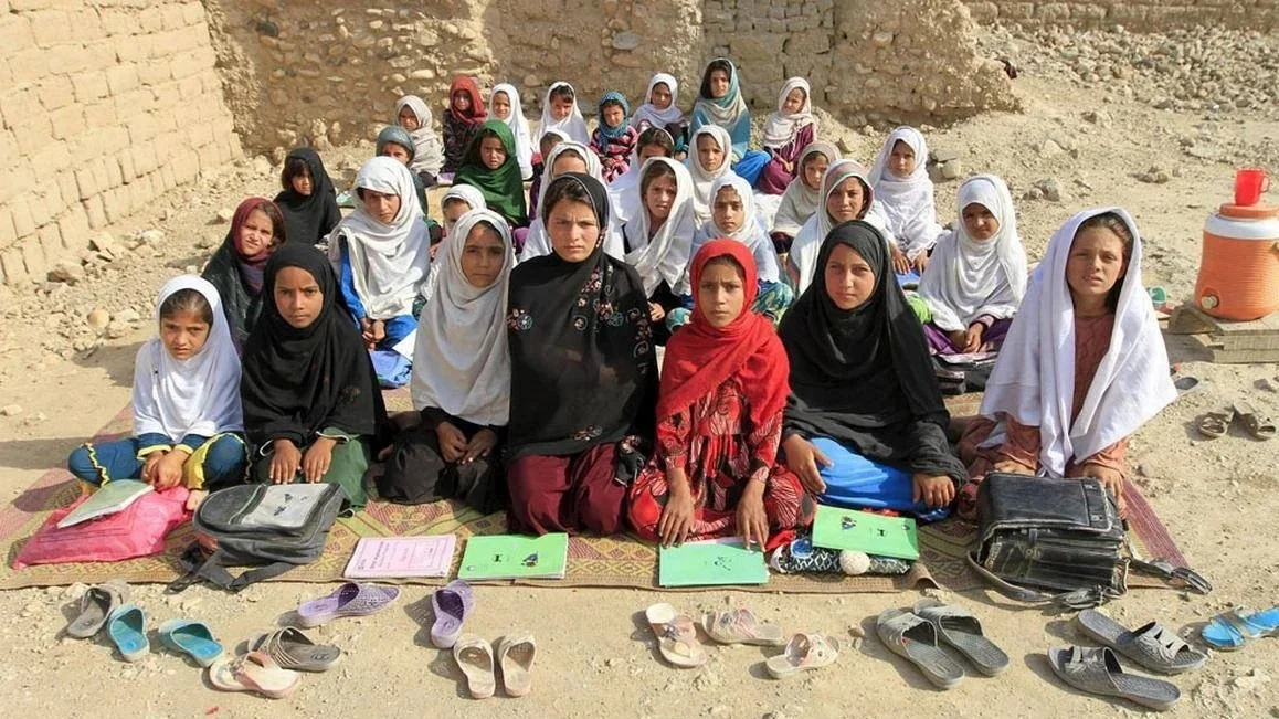 Group of children and women sitting on the ground outdoors, wearing traditional clothing, with shoes and notebooks in front of them.
