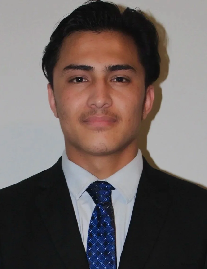 A young man with dark hair, wearing a white shirt, a patterned blue tie, and a black suit, standing against a light-colored background.