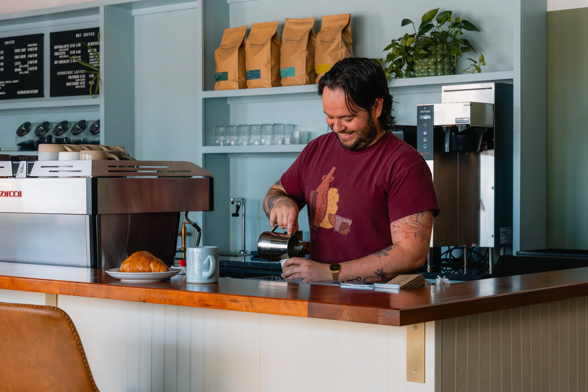 A man with tattoos and a beard smiling in a coffee shop, pouring milk into a cup, with a croissant on a plate in front of him, and coffee beans and a coffee machine in the background.