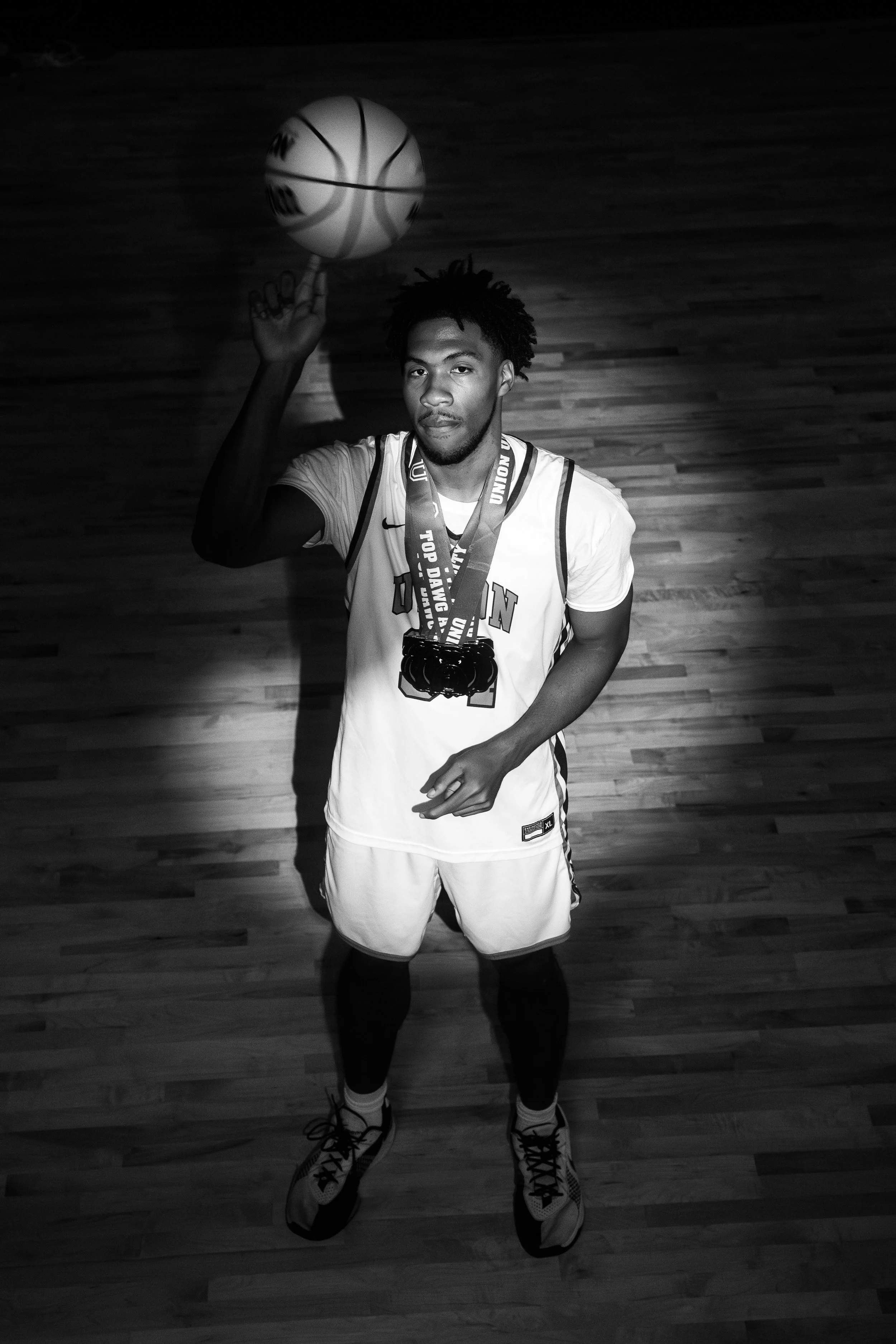 A young male basketball player standing on a hardwood court, wearing a sports uniform and medals, holding a basketball on his finger, and looking directly at the camera.
