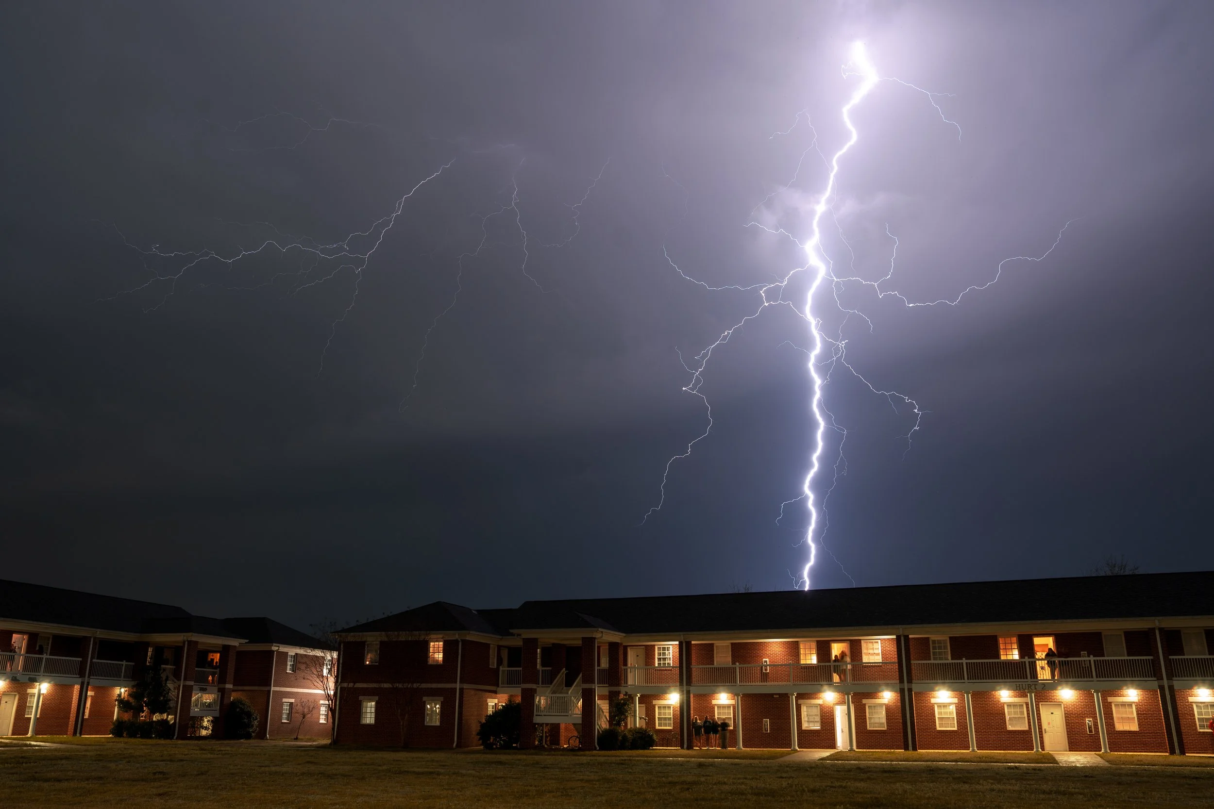 Nighttime scene with multiple lightning strikes in the sky above a two-story brick apartment complex with lit windows