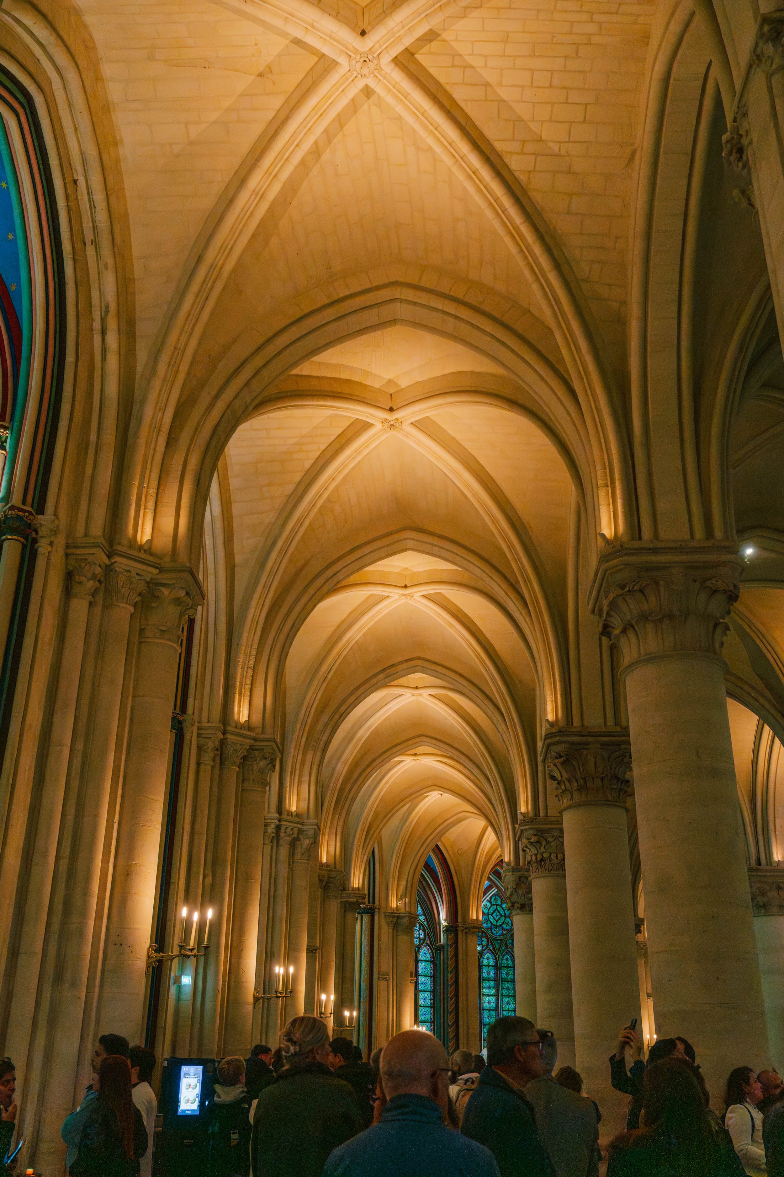Inside a large cathedral, with high vaulted ceilings and stained glass windows, a crowd of people is gathered, some looking at their phones and taking pictures.