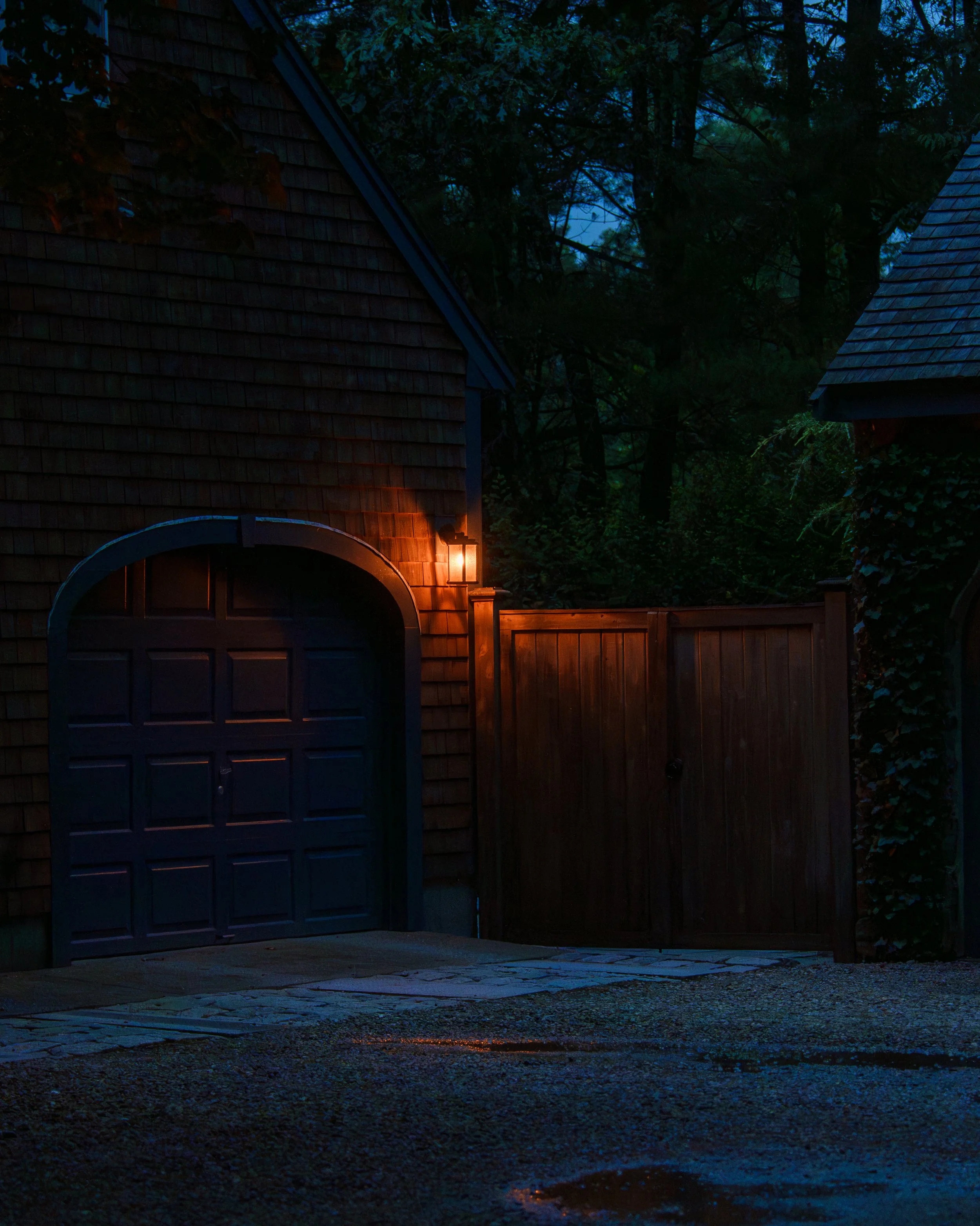 Nighttime scene of a house with a wooden gate and an arched garage door, illuminated by a wall-mounted lantern, surrounded by trees and shadows.