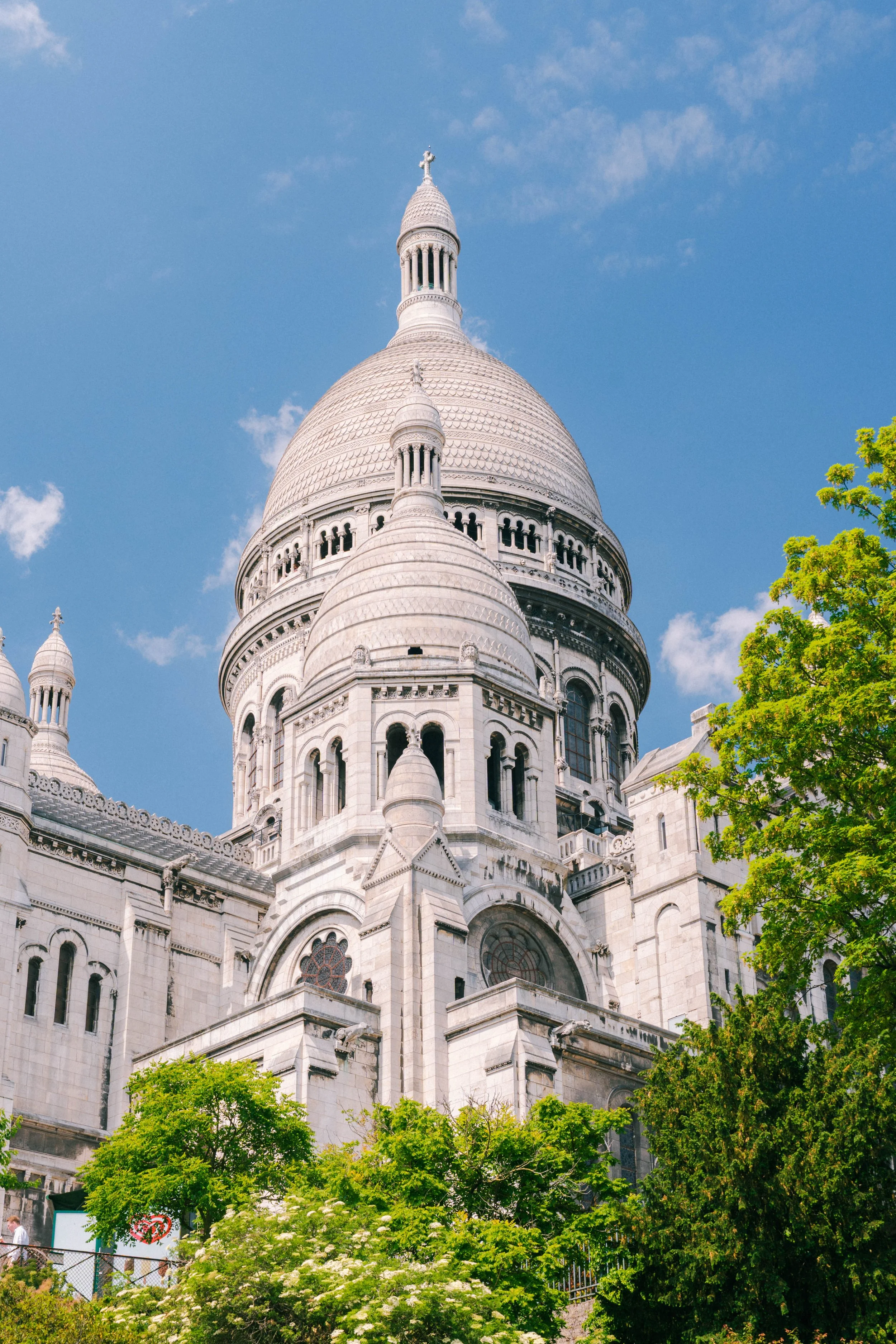 The Sacré-Cœur Basilica, a white domed cathedral, in Montmartre, Paris, France, with trees and a blue sky in the foreground.