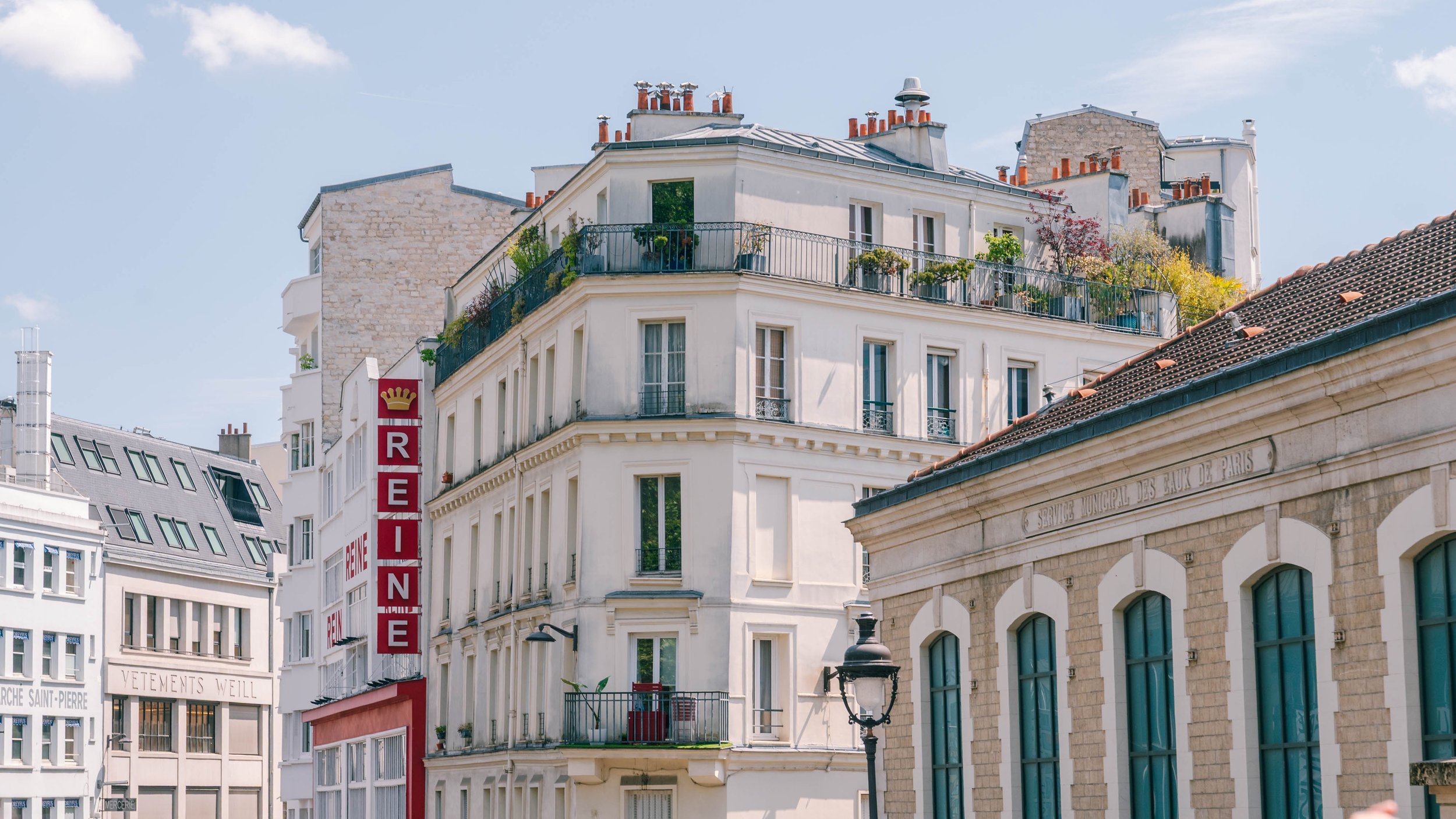 Street view of Paris buildings with a prominent white building featuring a rooftop garden, a red and white hotel sign, and a stone building with an inscription in French.