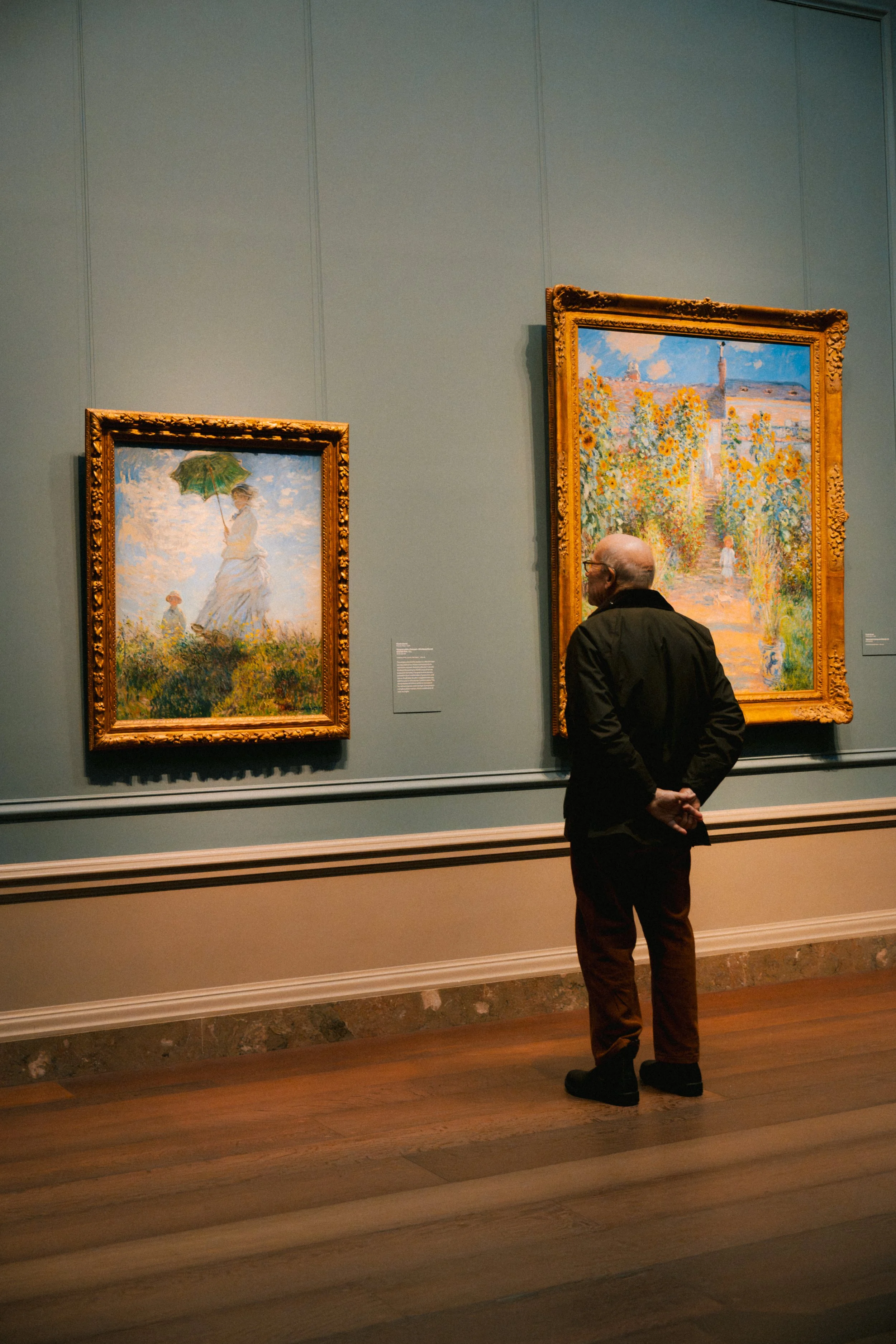 A man with glasses and a beard stands with his hands behind his back, observing impressionist paintings of outdoor scenes in a museum.
