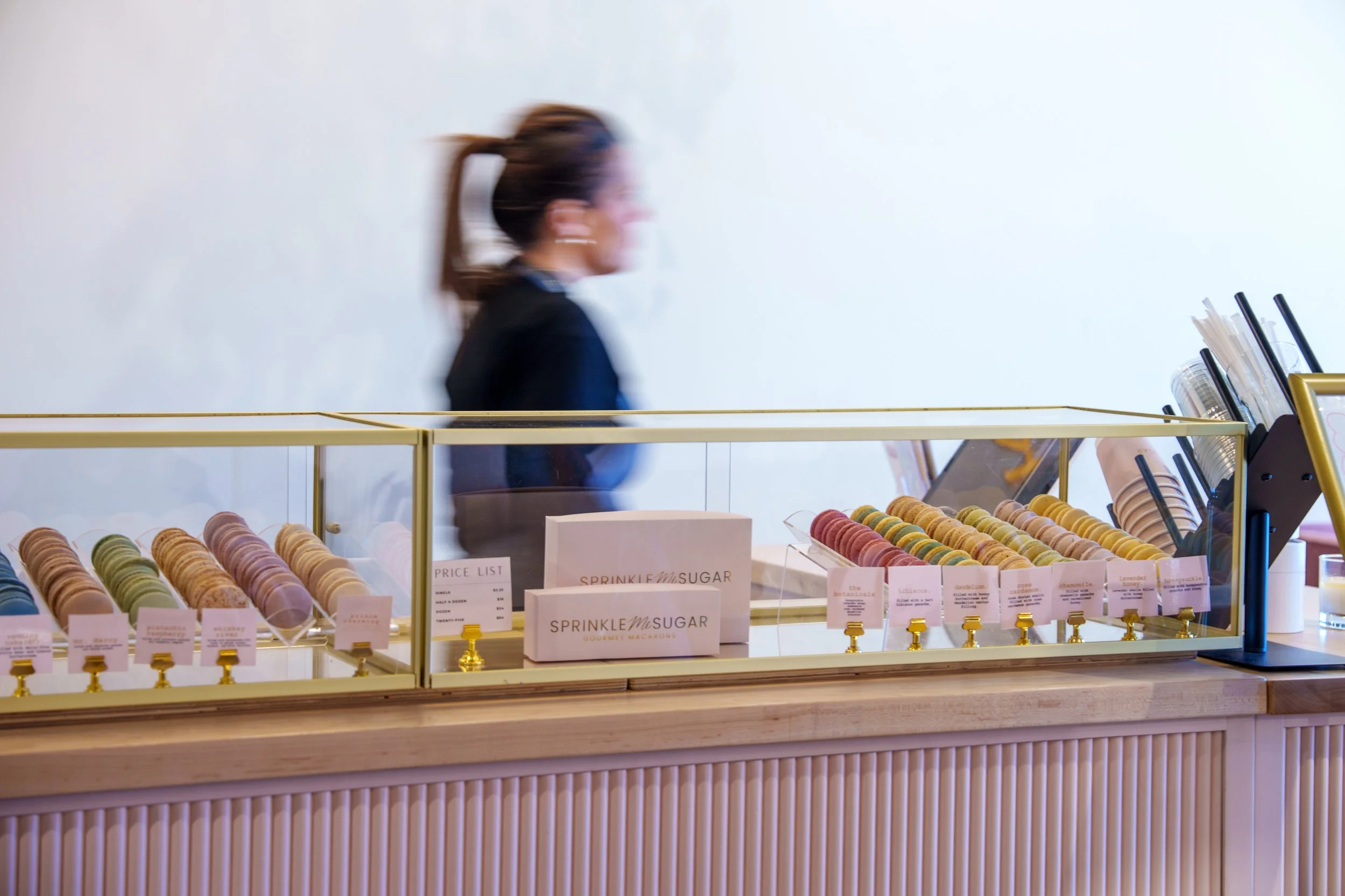 Blurred image of a woman walking past a display of colorful macarons at a bakery or pastry shop.