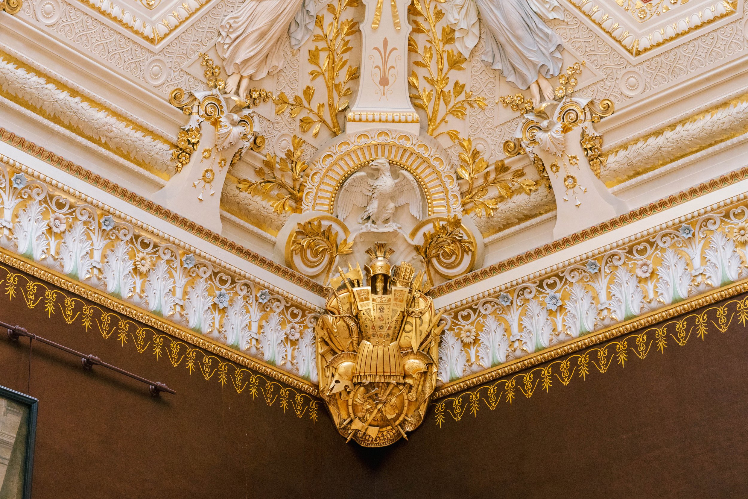 Decorative ceiling with ornate gold and white detailing, featuring a carved eagle and figure sculptures, surrounded by intricate floral and geometric patterns.