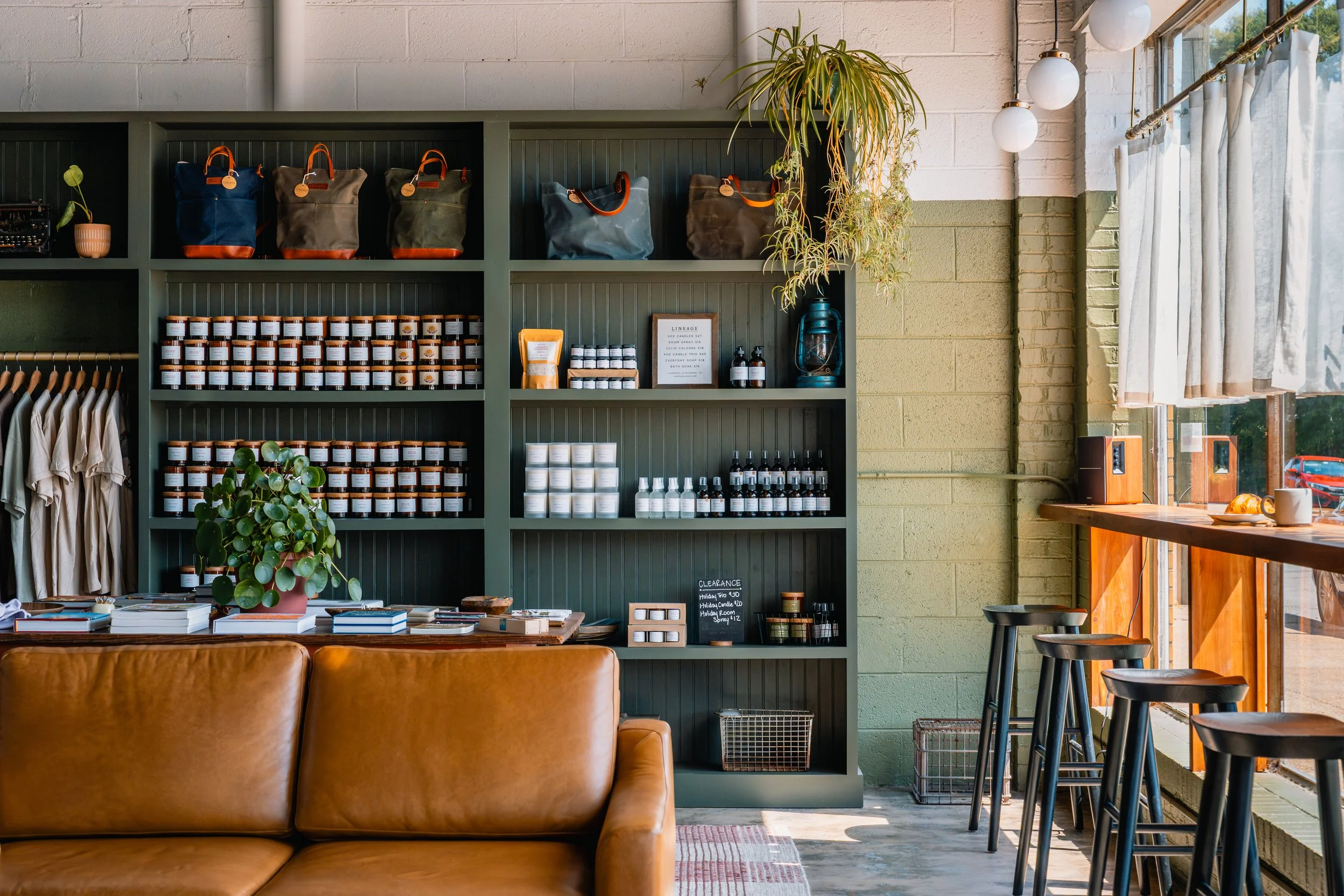 Interior of a shop or café with a tan leather sofa in the foreground, a gray shelving unit with products, plants, and framed menu, and a large window with white curtains letting in natural light.
