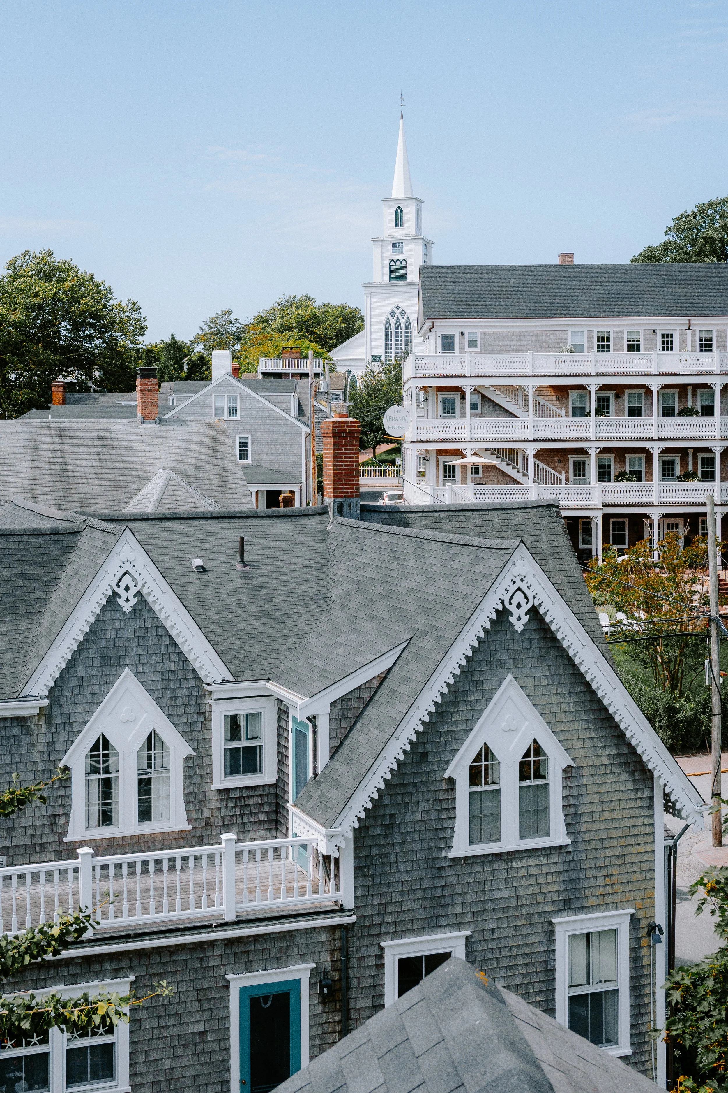 View of residential houses with grey shingle siding and white trim, including decorative gable ends, dormer windows, and a balcony; a white church with a tall steeple is visible in the background under a blue sky.