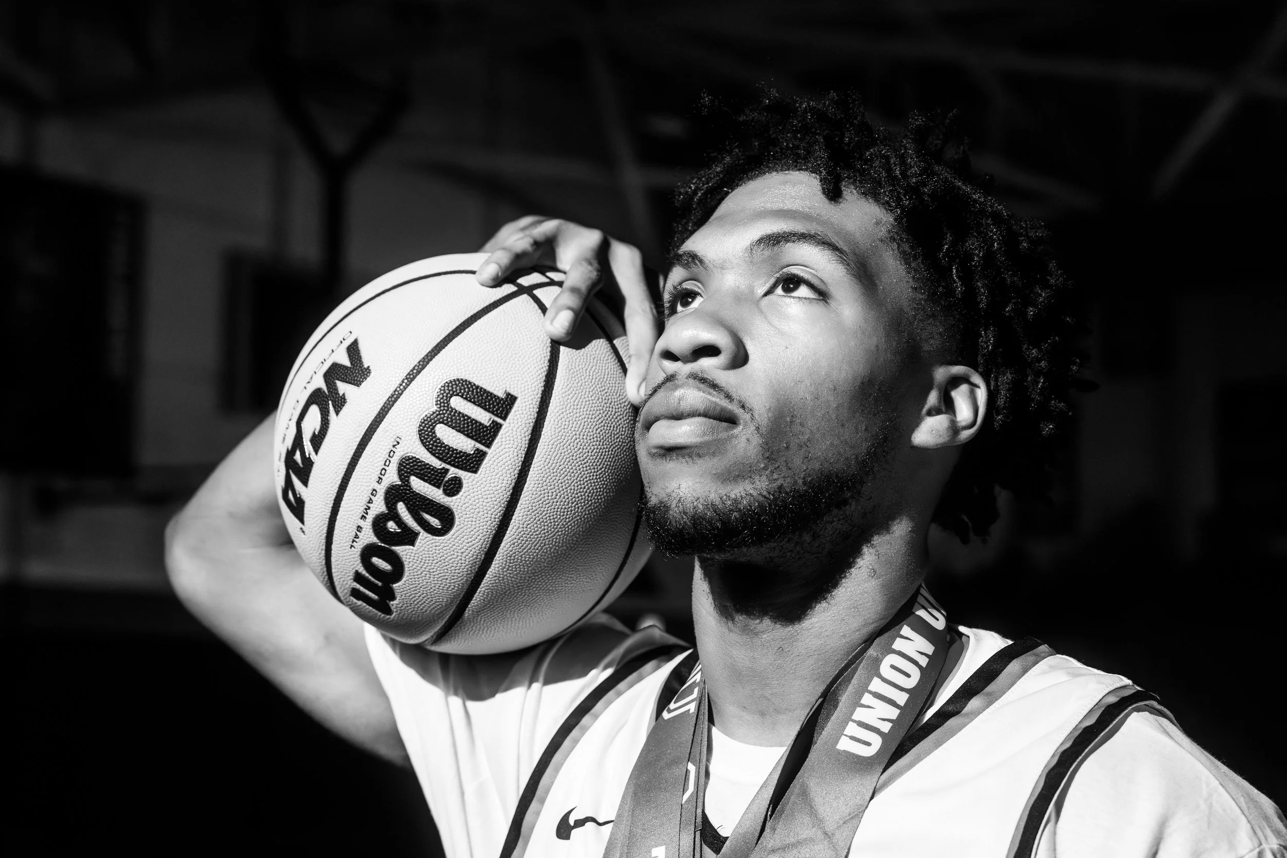 A young man with curly hair and a goatee holding a basketball close to his face, wearing a sports jersey and a medal around his neck.