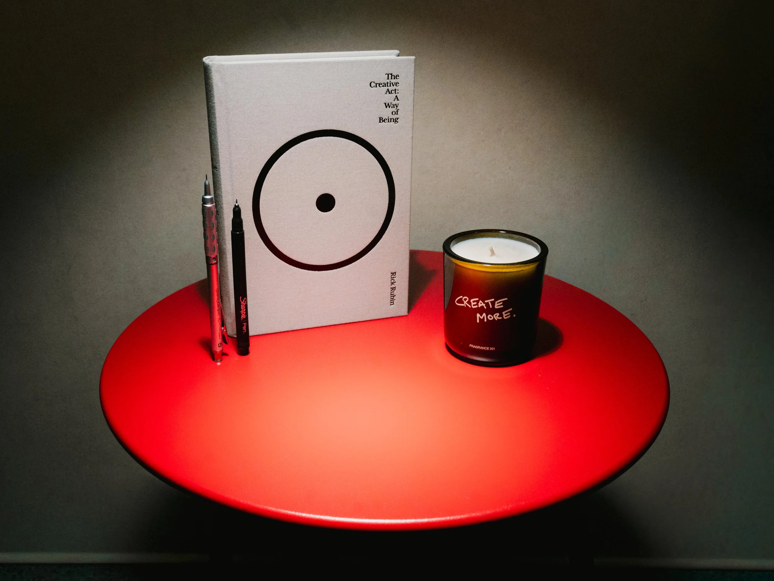 A red circular table with a white book, two pens, a black candle with the words 'Create More,' and a small white candle on top.