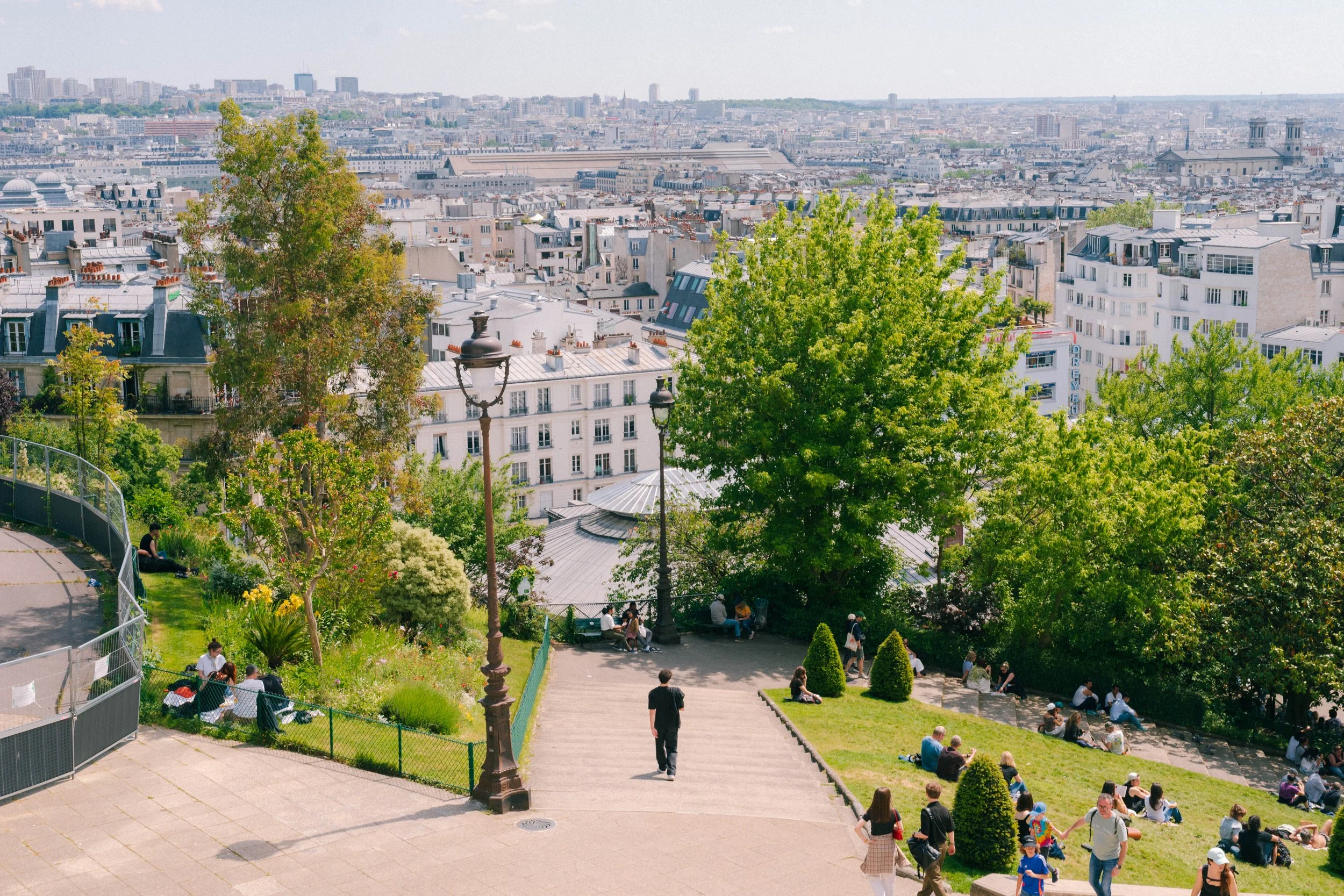 A park on a hill overlooking a cityscape with buildings and trees, people sitting and walking, sunny day.