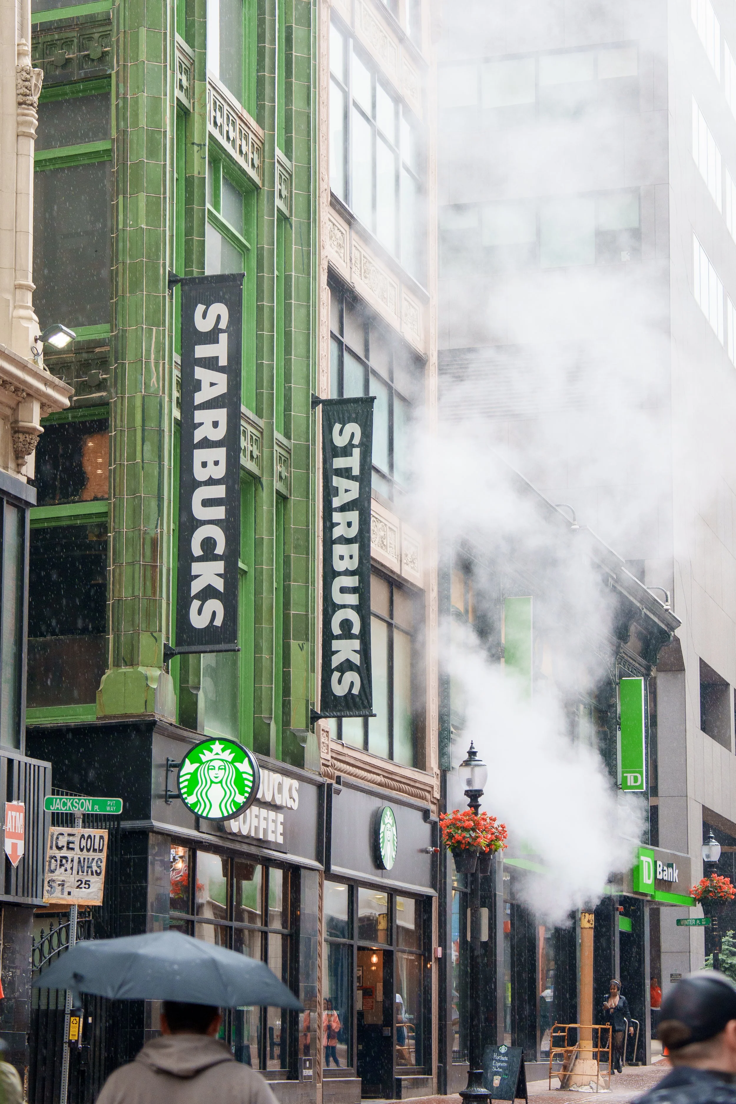Street scene with a Starbucks coffee shop, green building with black banners reading 'Starbucks', a person holding an umbrella, and steam rising from underground vents.