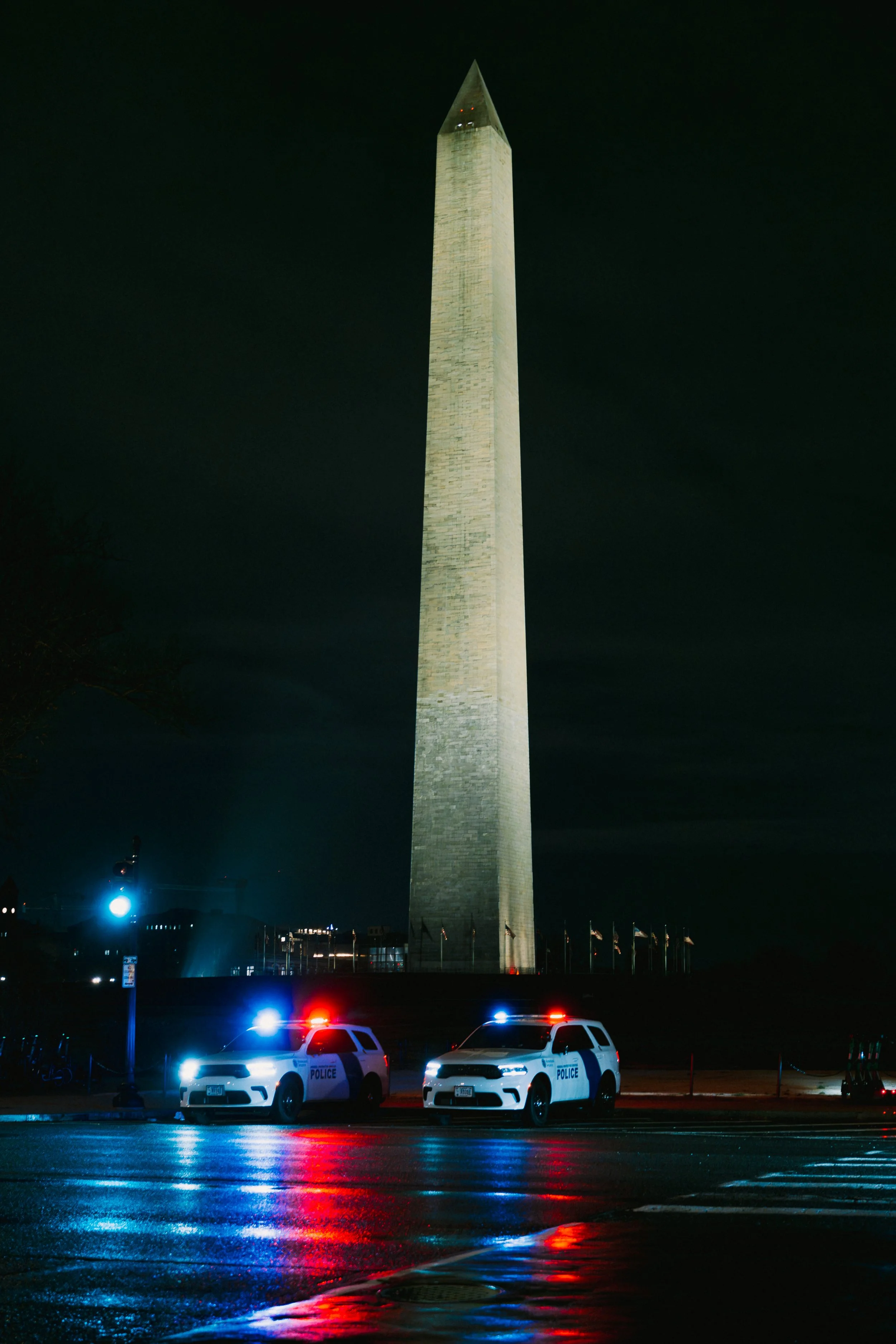 Night view of the Washington Monument illuminated in Washington D.C., with two police cars with flashing lights in the foreground and reflections on the wet street.