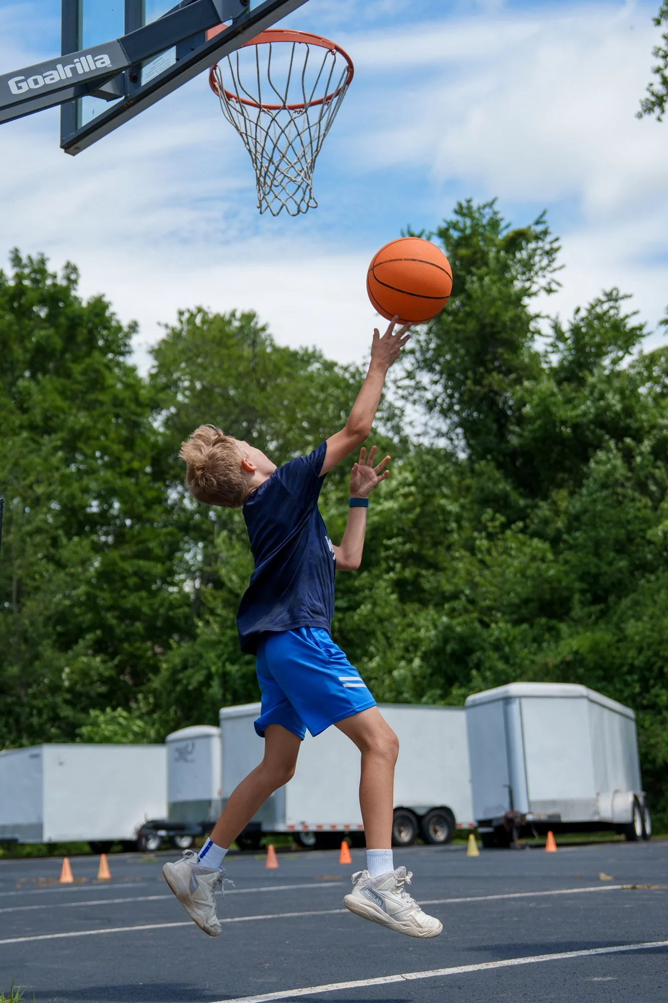 Young boy playing basketball outdoors on a parking lot, jumping to shoot the ball toward the hoop, with green trees and trailers in the background.