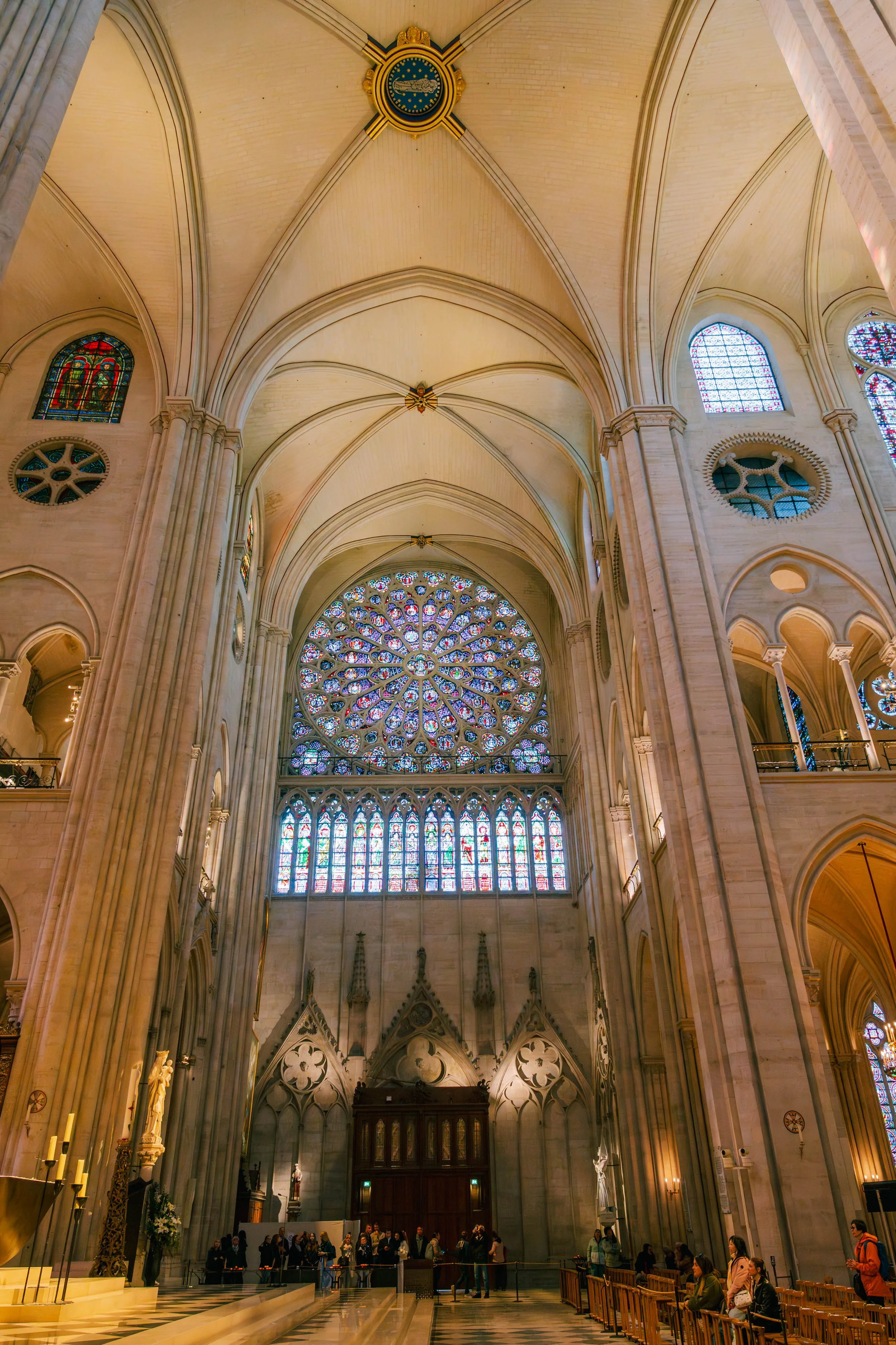 Interior of a grand Gothic cathedral with high vaulted ceilings, stained glass windows, and a large rose window. People are seated and standing underneath.