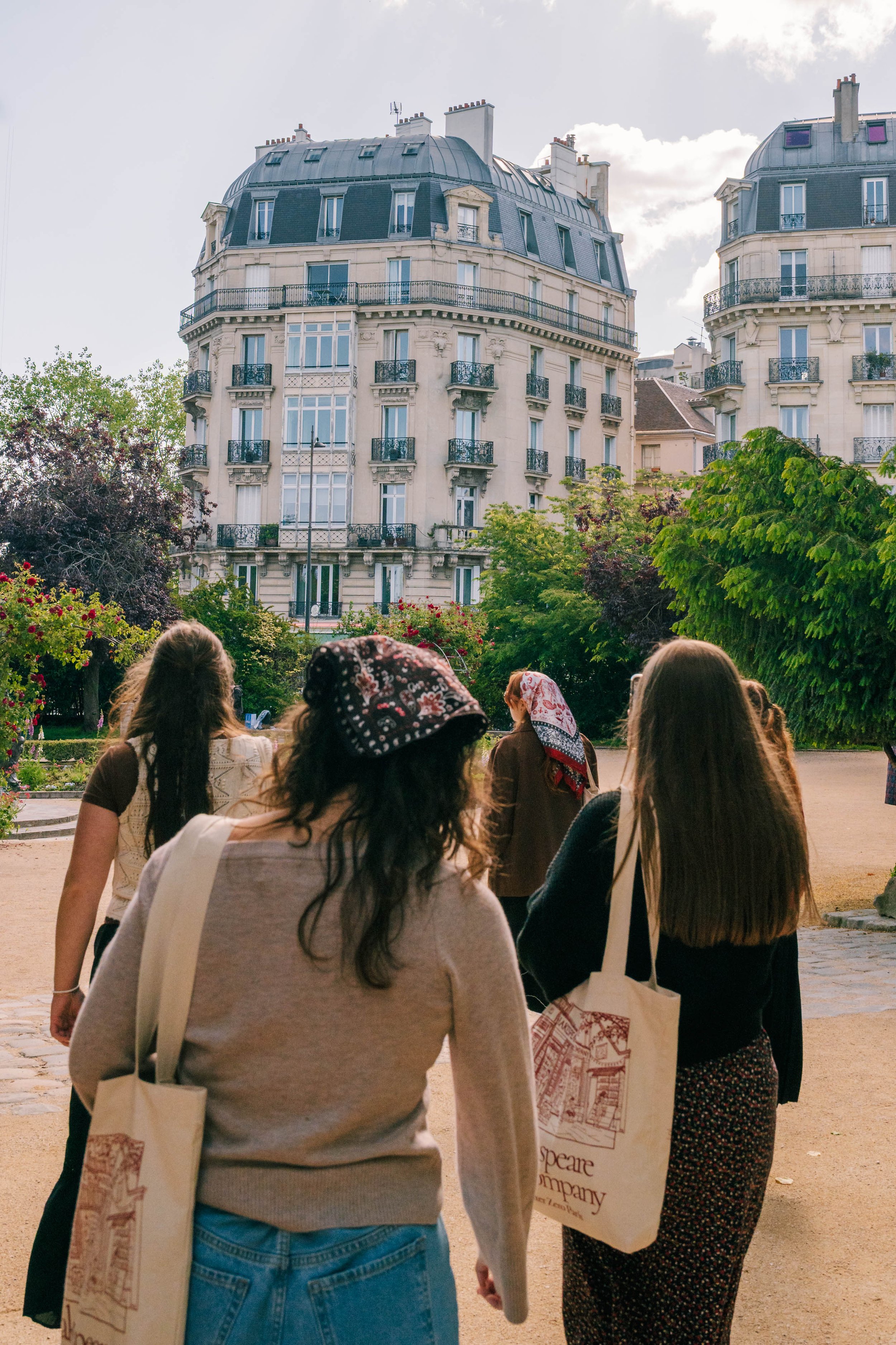 Group of women walking outdoors in front of historic Parisian-style buildings with multiple stories and mansard roofs, surrounded by trees and plants.