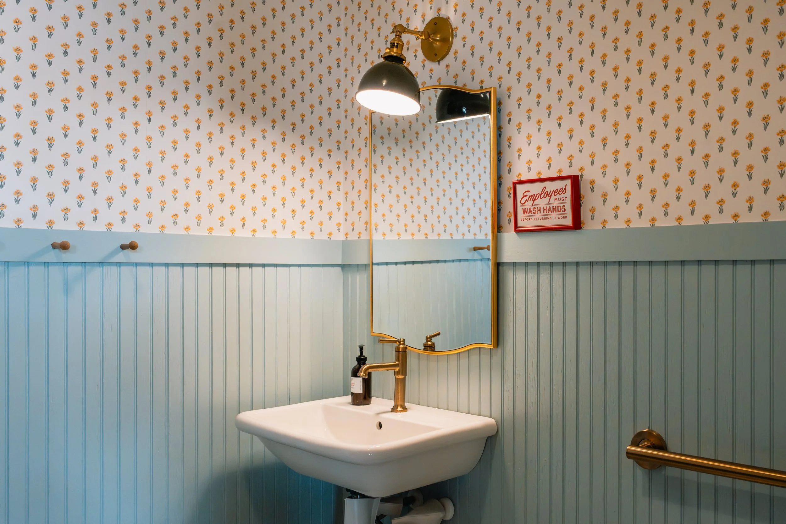 A bathroom with floral wallpaper, a wall-mounted light fixture, a mirror, a small framed sign, a faucet, and a sink.