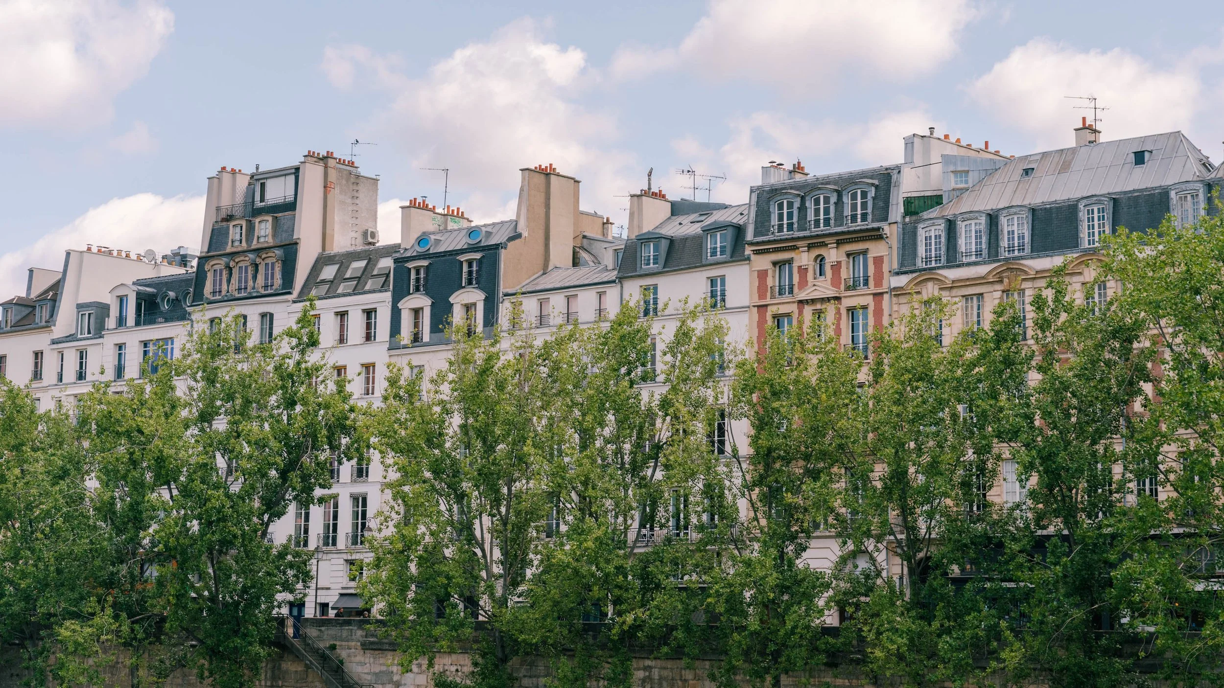 Parisian apartment buildings with gray roofs and dormer windows, with green trees in the foreground and a partly cloudy sky in the background.