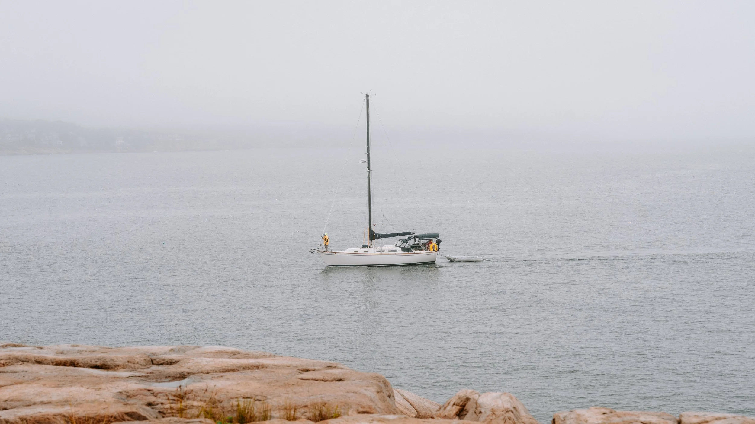 A sailboat with a single mast and a small boat being towed is seen near a rocky shoreline on a foggy day over calm water.