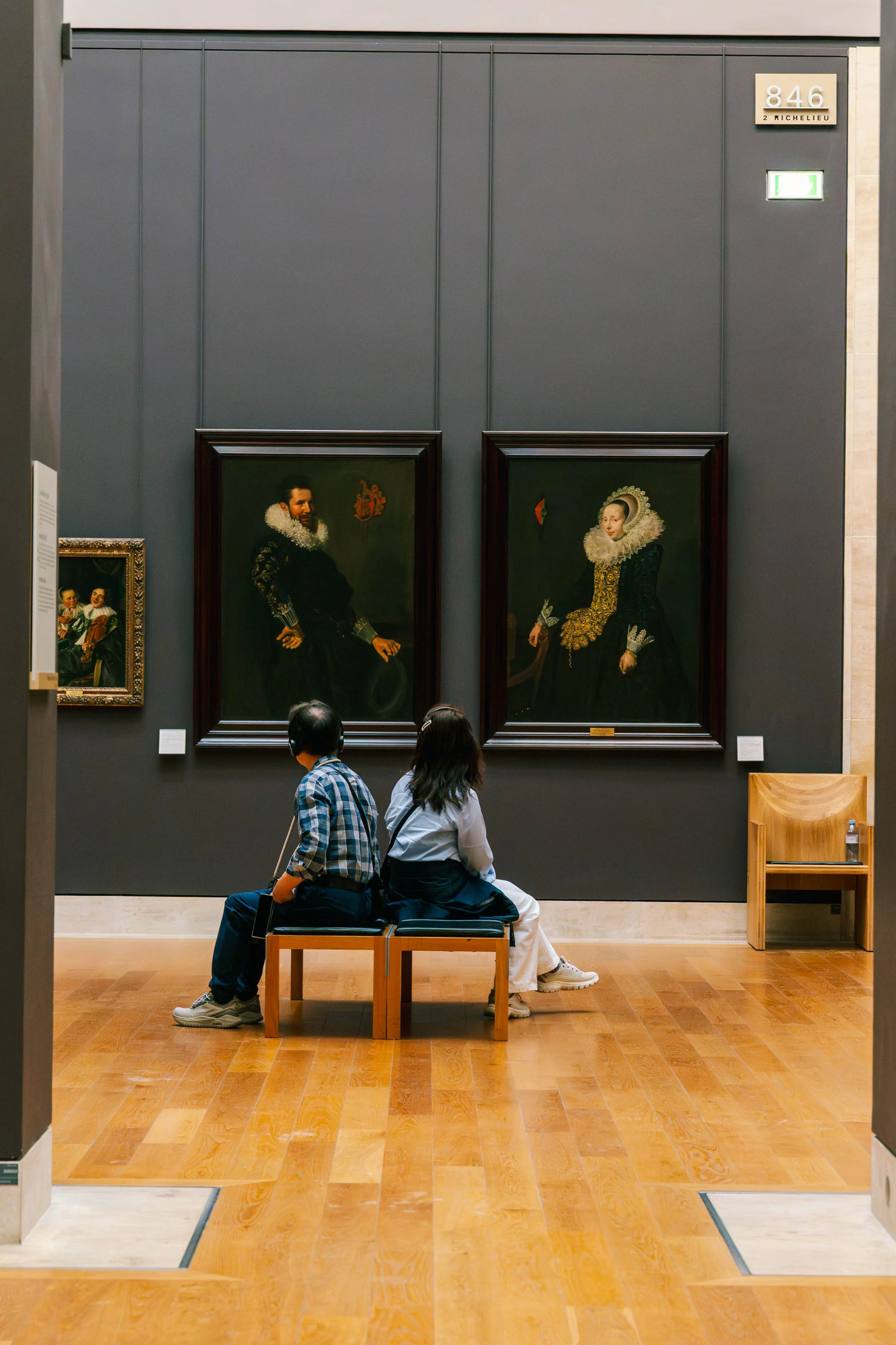 Inside an art gallery or museum, two people are sitting on a bench in front of large portrait paintings hanging on a dark gray wall. The portraits depict a man and a woman in historical clothing. There are two small plaques below each painting, and a