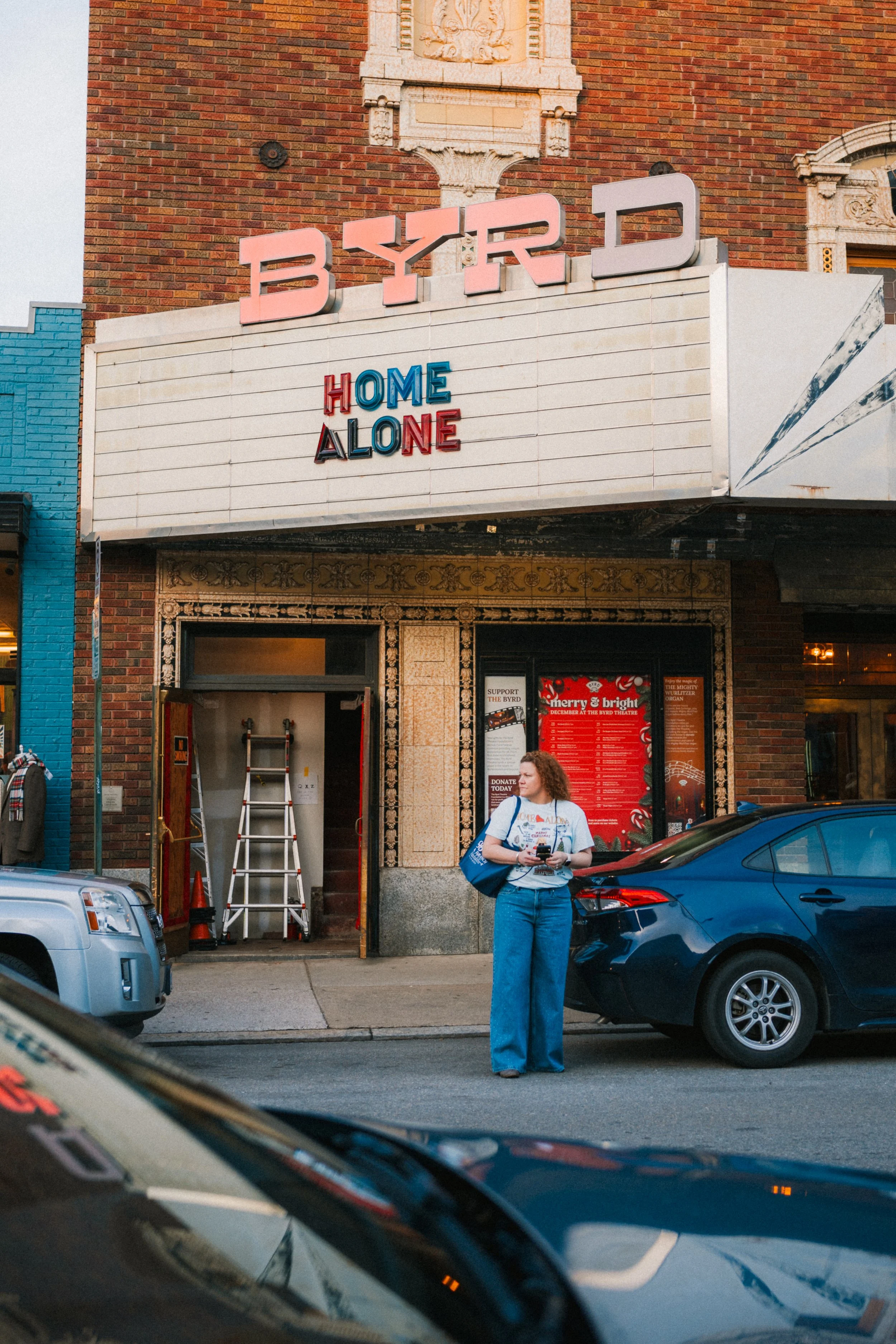 A woman with curly hair standing on the sidewalk in front of the Byrd Theatre, holding a smartphone and looking to the side. The theatre's marquee displays the title 'Home Alone.' Two parked cars are visible, and a ladder is inside an open door of th