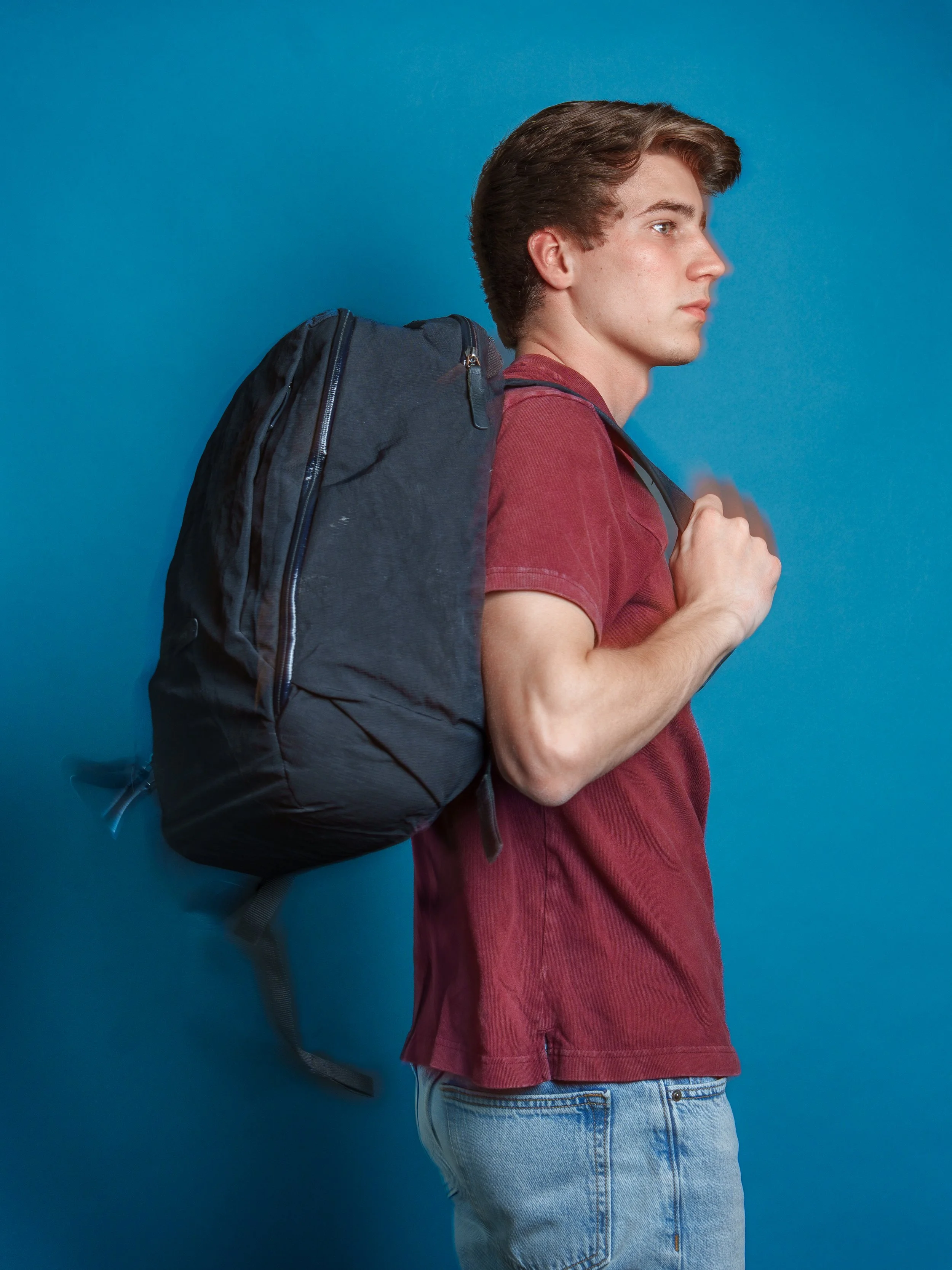 A young man with brown hair, wearing a red t-shirt and light blue jeans, is standing against a blue background, carrying a large dark backpack on his back, holding the shoulder straps with both hands, in a profile view.