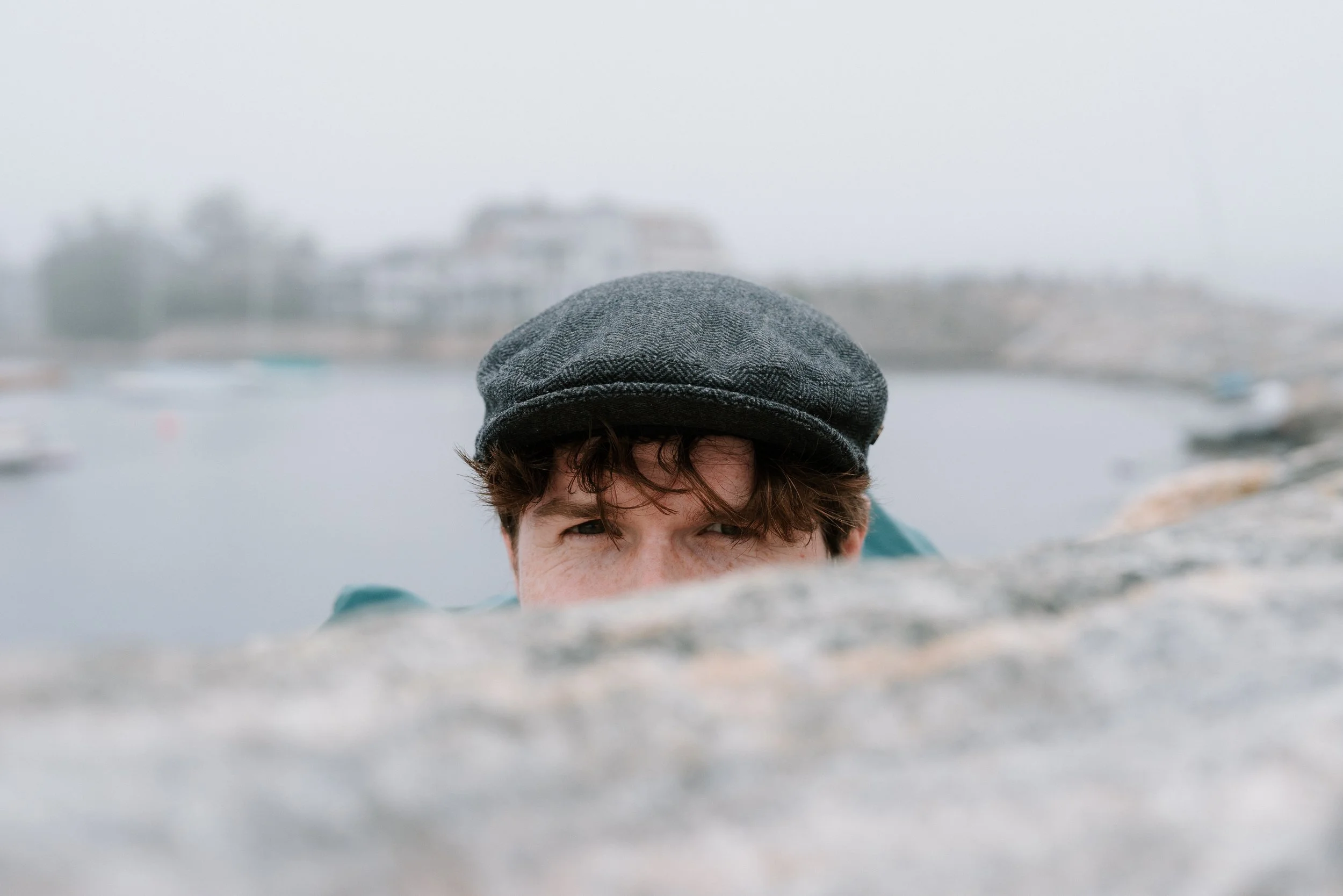 Person peeking over a stone wall, wearing a gray cap, with water and buildings in the background.