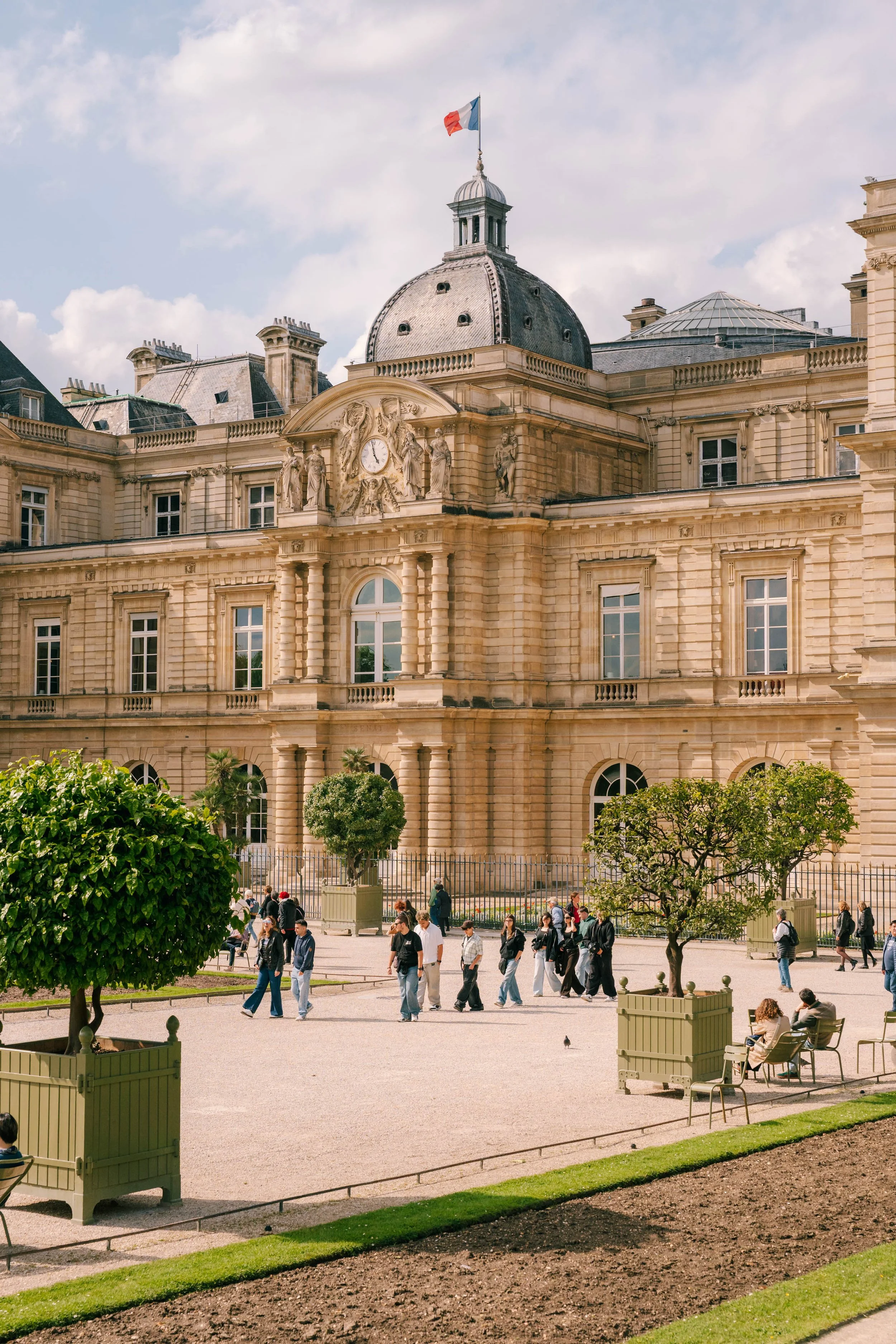 A historic beige stone building with a domed roof, a clock, and French flag on top. People walking and sitting in a park area with trees and benches in front.