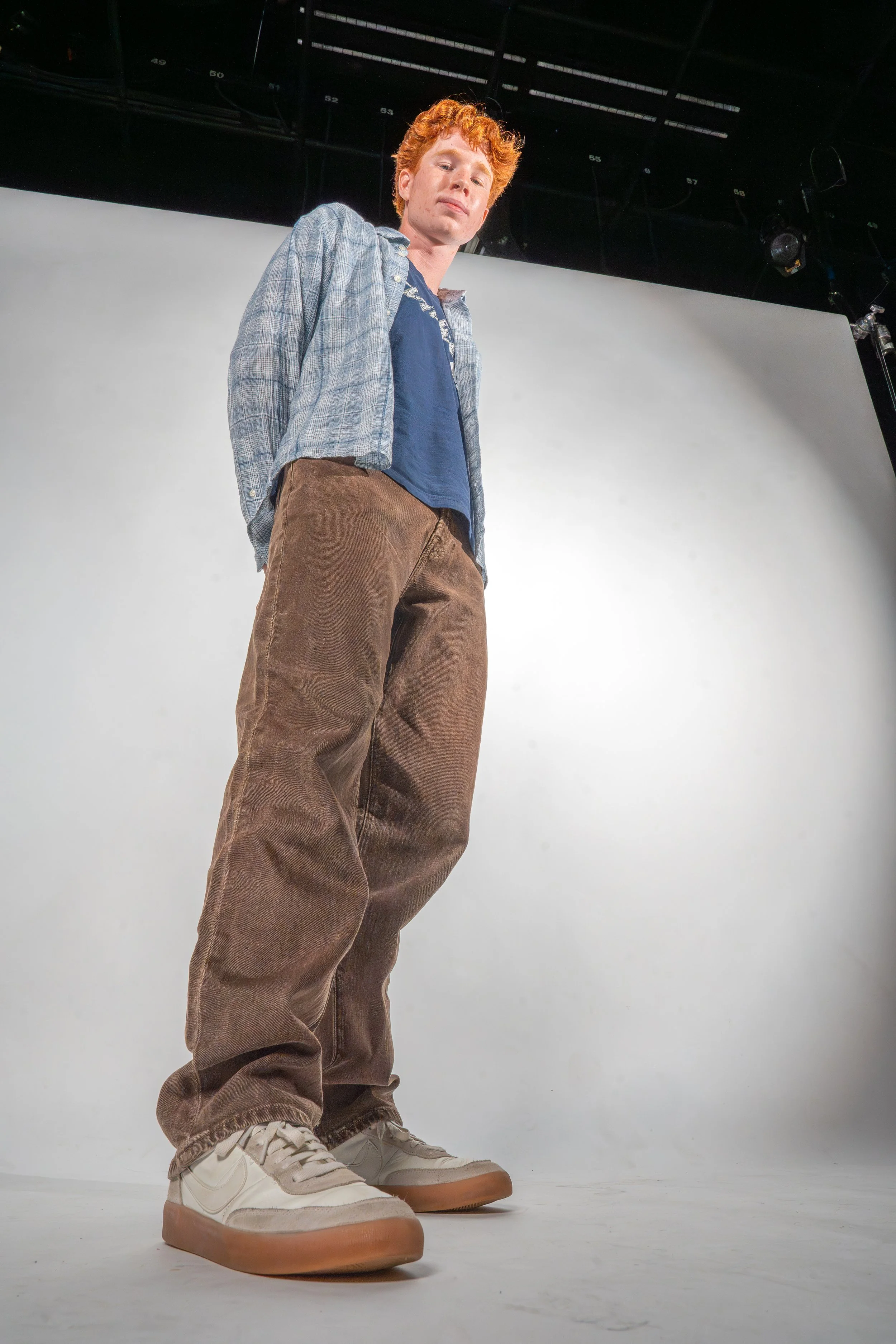 Young man with curly red hair wearing a blue T-shirt, plaid shirt, brown pants, and white sneakers standing against a plain white backdrop in a photography studio.