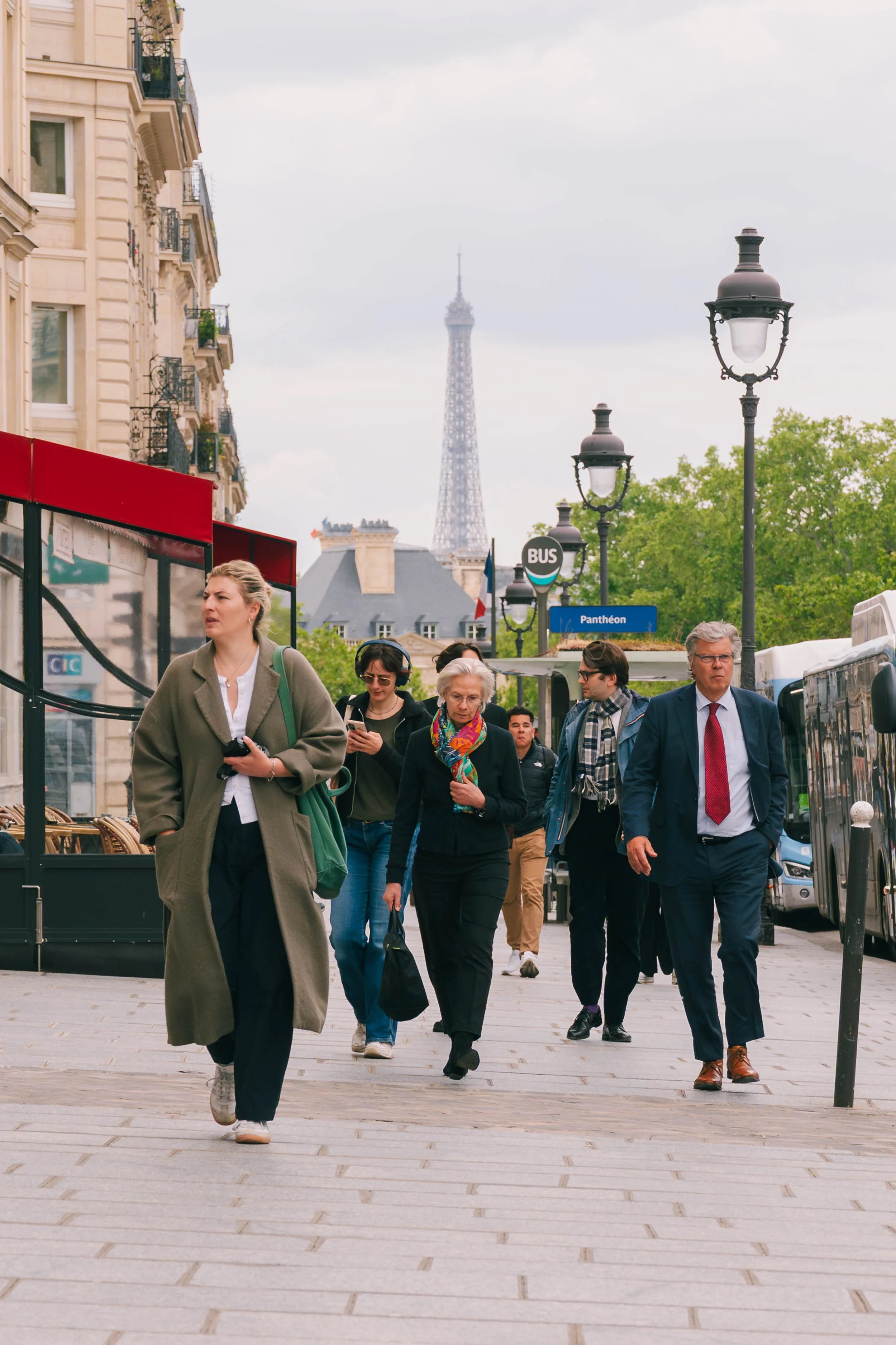 A group of diverse people walking on a Paris city street with the Eiffel Tower in the background. The street has traditional Parisian street lamps and a bus stop sign that reads 'Pantheon'.