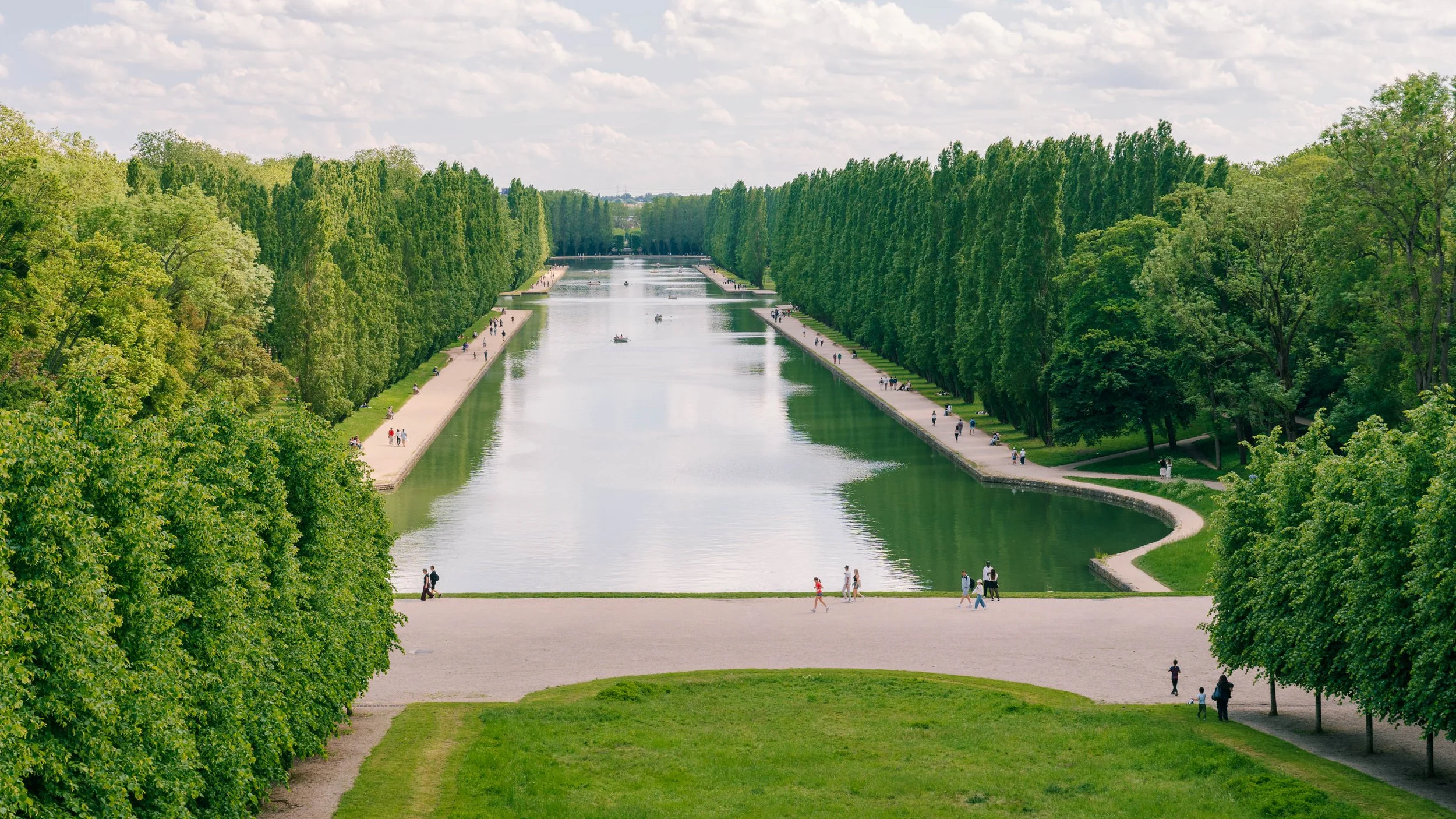 A wide view of a large, rectangular green park with a canal running through the middle, lined with lush green trees on both sides, and people walking along the paths.