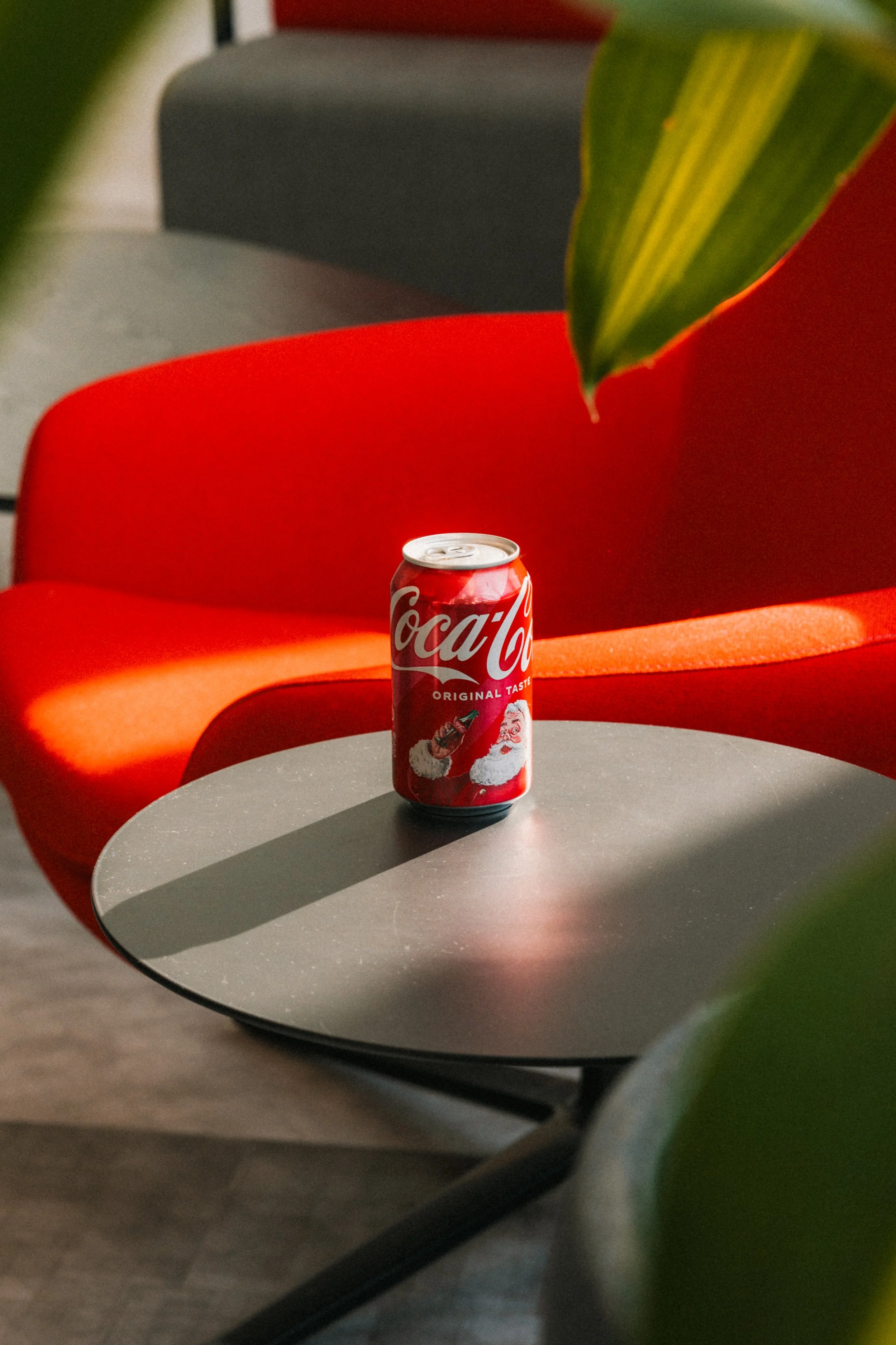 A can of Coca-Cola with a holiday Santa Claus design sits on a gray table, with red and gray chairs and top of a leafy plant in the foreground.
