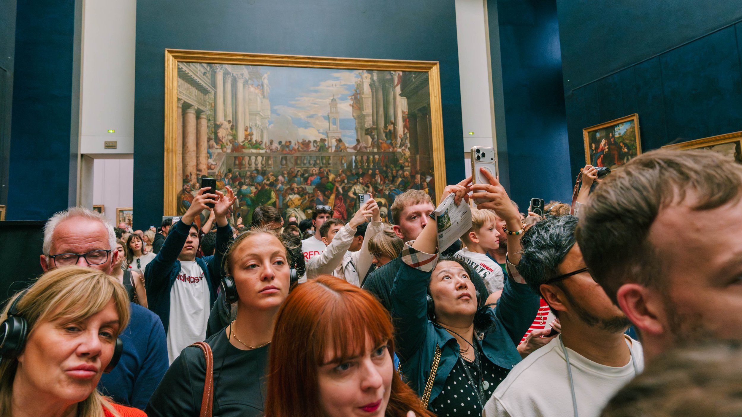 Crowd of people inside an art gallery, many taking photos of a large classical painting on the wall.