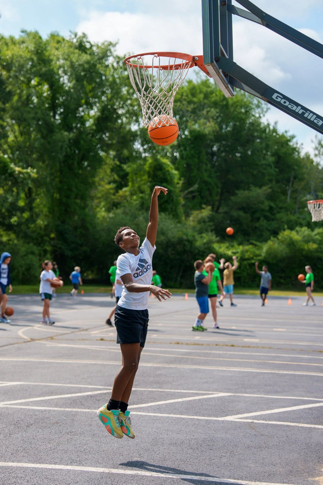 A young boy in a white Adidas shirt and dark shorts jumping to make a basketball shot on an outdoor court, with other children practicing basketball in the background.