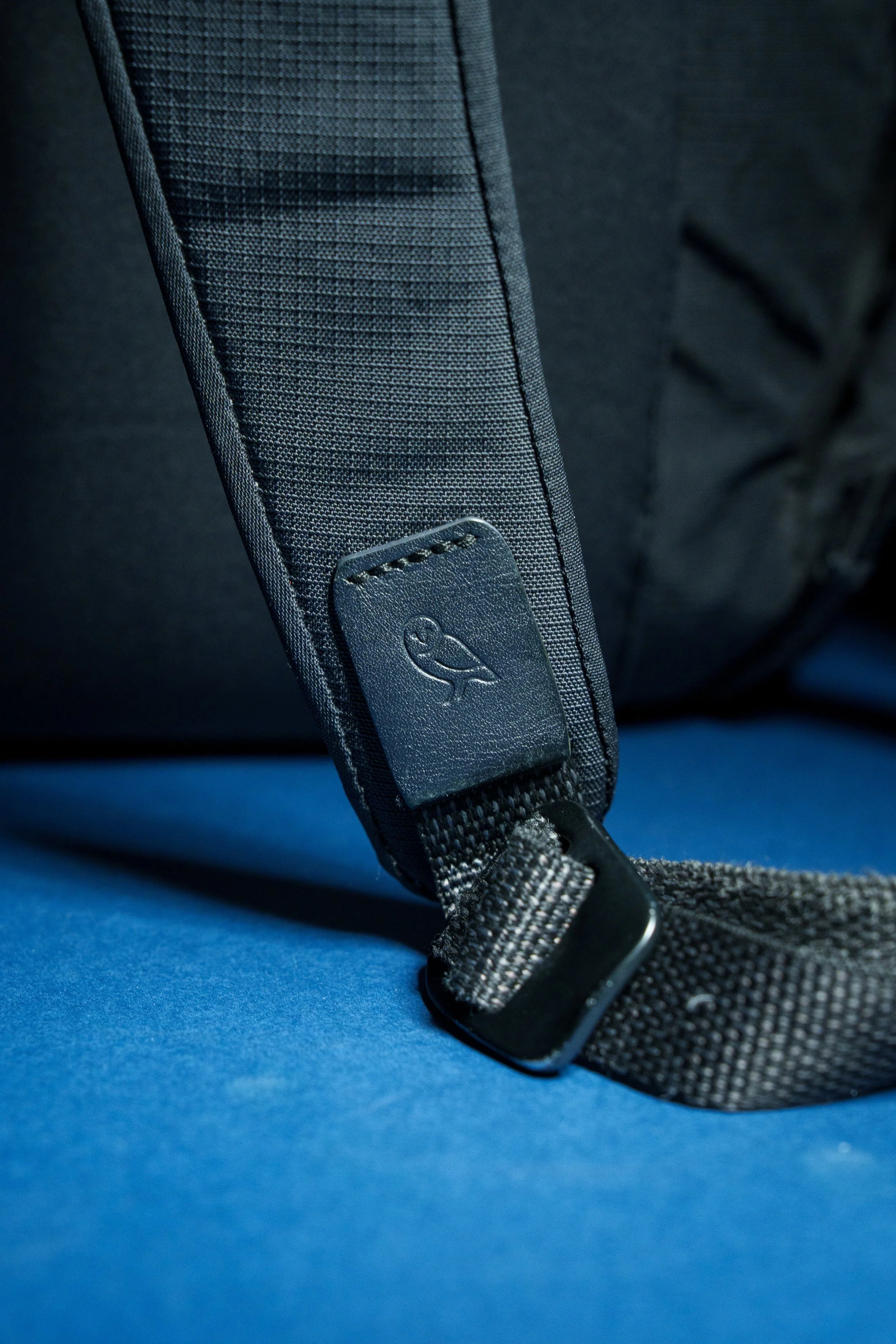 Close-up of a dark gray backpack strap with a small leather detail and metal buckle on a blue surface.