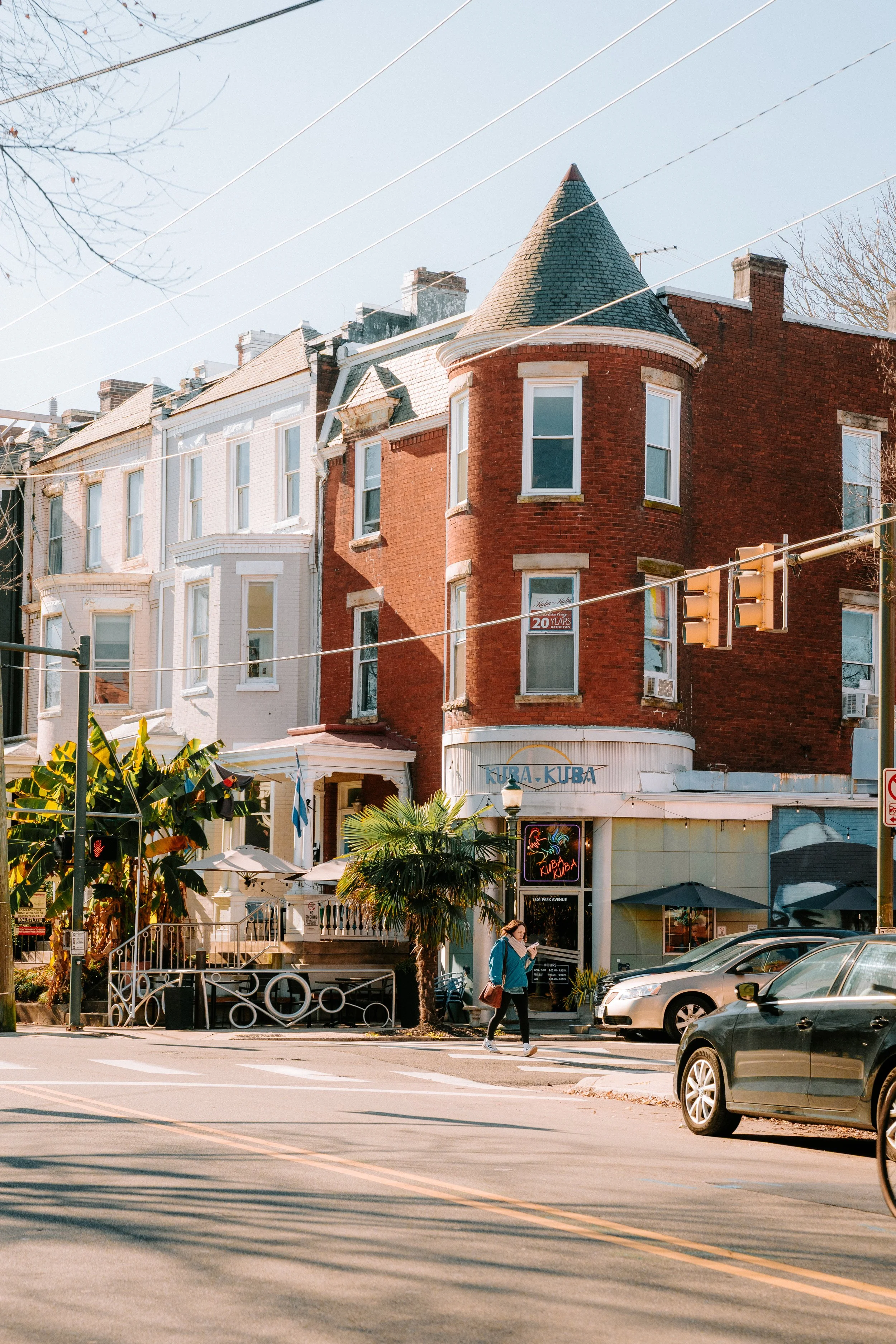 A street scene featuring three row houses, one red brick with a turret and two white with bay windows, and a storefront called 'Kura Kura' with outdoor seating and palm trees. Pedestrians and cars are visible.