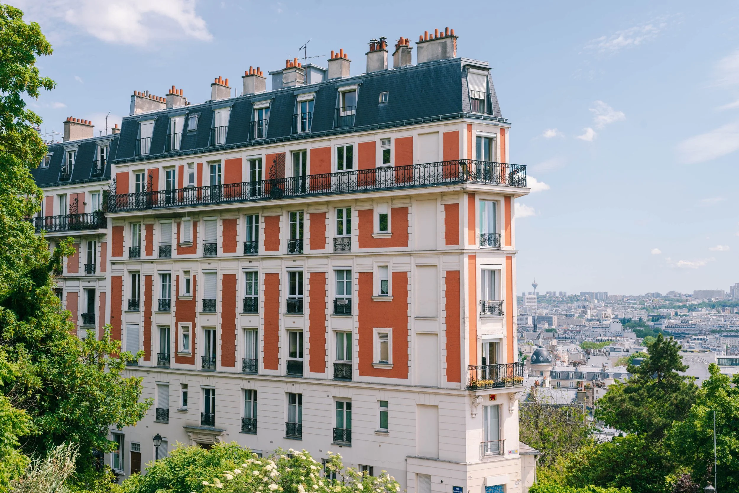 A multi-story residential building with orange and white exterior walls, several balconies with black railings, and a blue mansard roof with multiple chimneys. The building is surrounded by green trees and there is a city skyline with a tower in the 