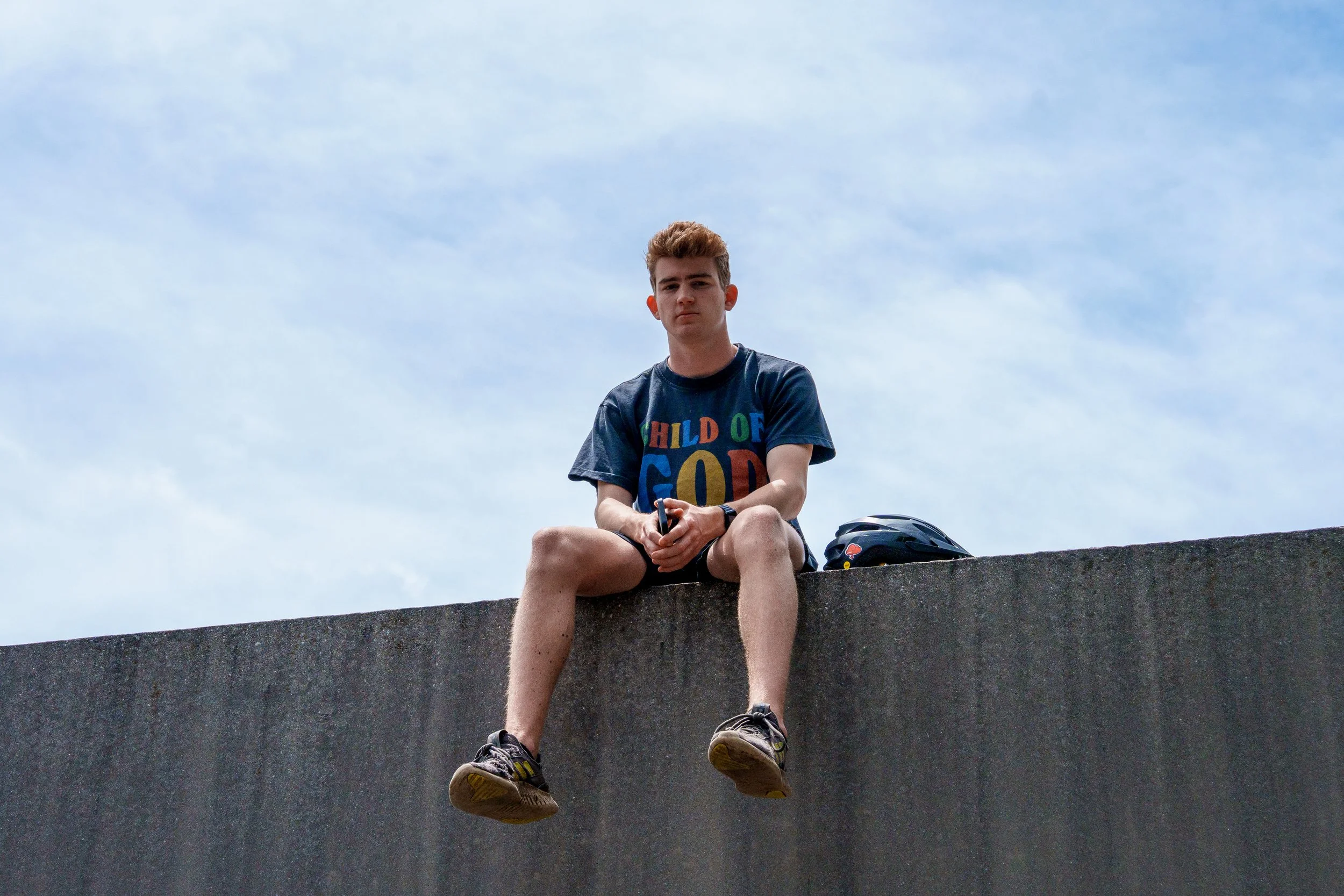 A young man sitting on a concrete ledge with a helmet beside him, wearing a graphic T-shirt and shorts, against a partly cloudy sky.