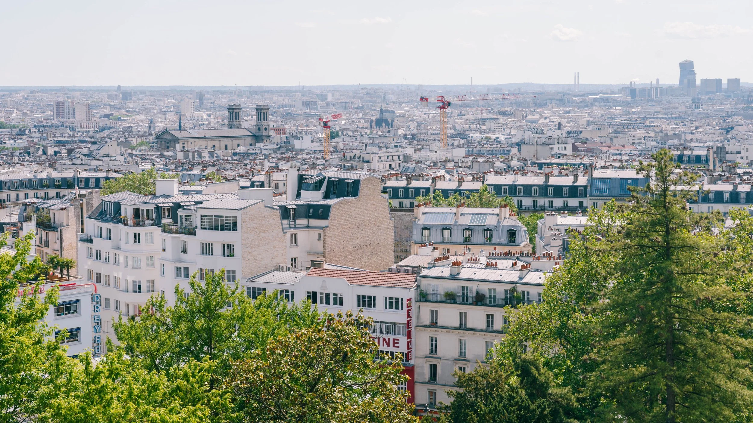 View of Paris cityscape with rooftops, trees in the foreground, and cranes in the distance under a partly cloudy sky.