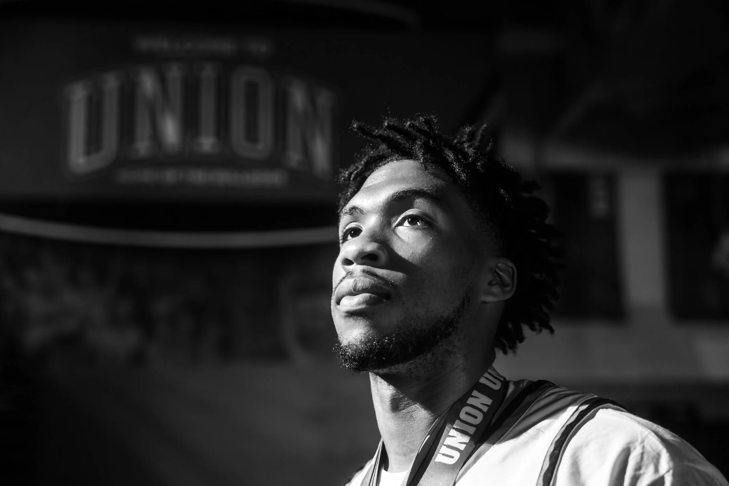 Black and white photo of a young man with dreadlocks looking up, wearing a sports jersey and a lanyard around his neck.