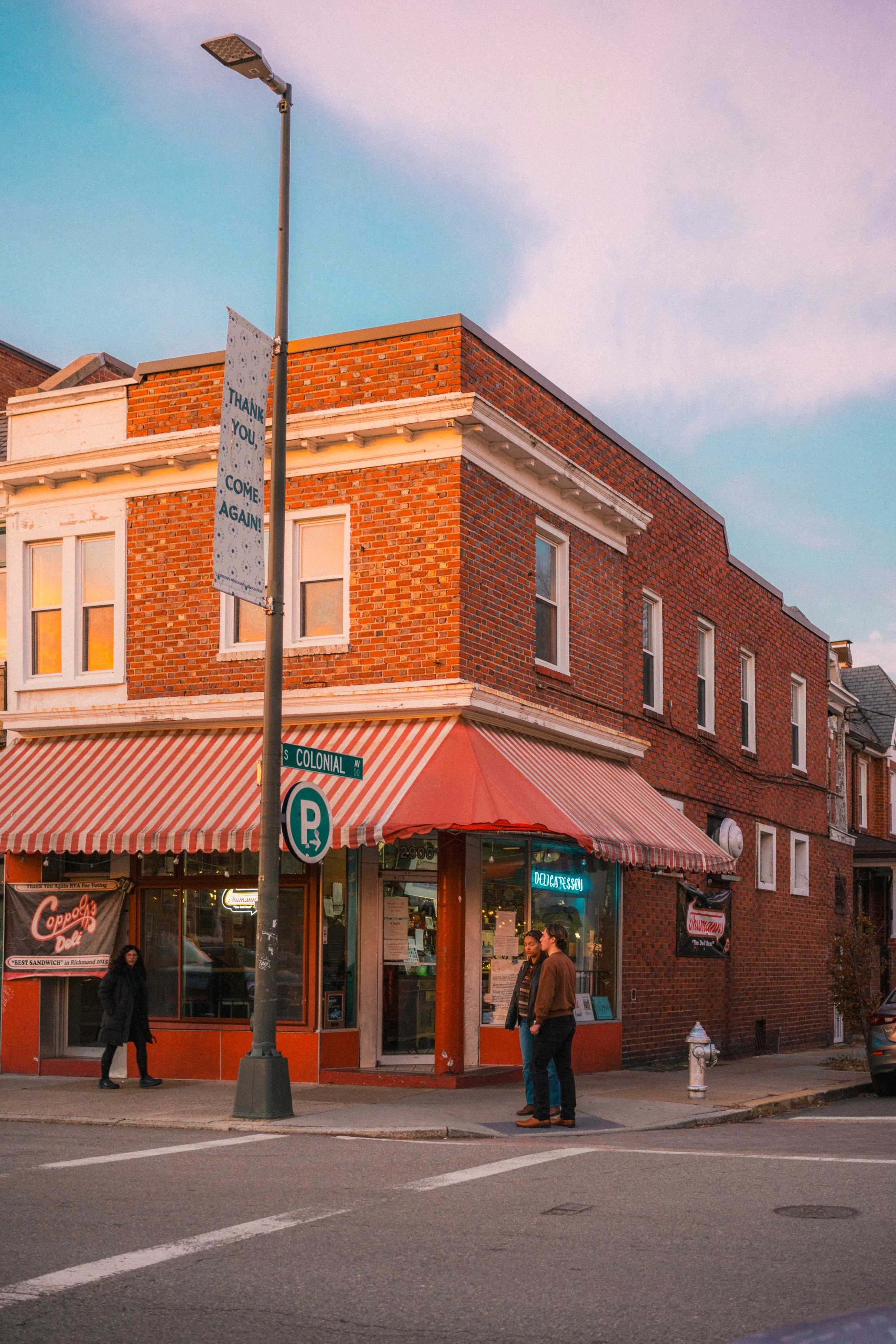 A corner of a brick building with a storefront, striped red and white awning, street signs, and people walking on the sidewalk during sunset.