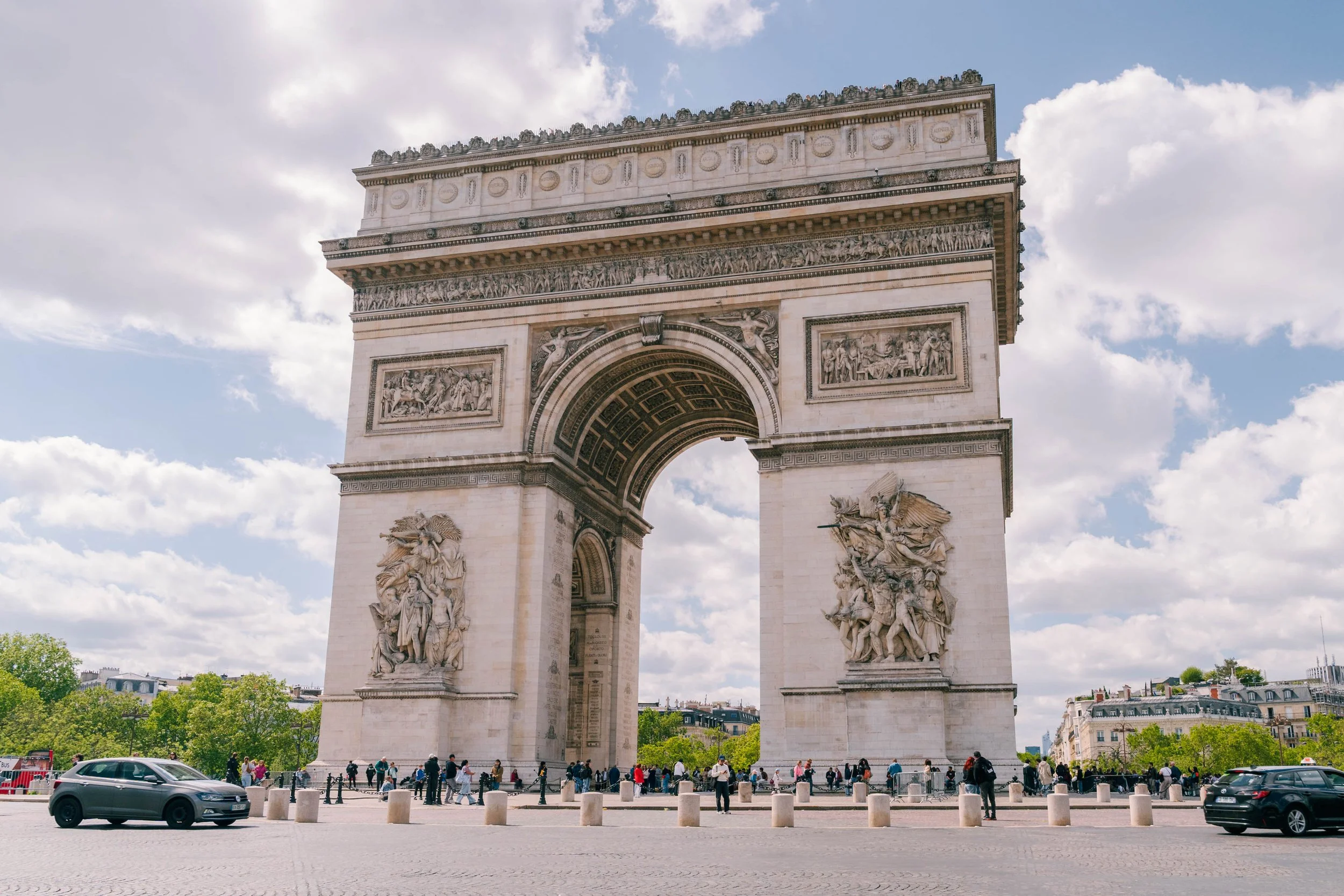 Arc de Triomphe in Paris under a partly cloudy sky with people walking and cars nearby.