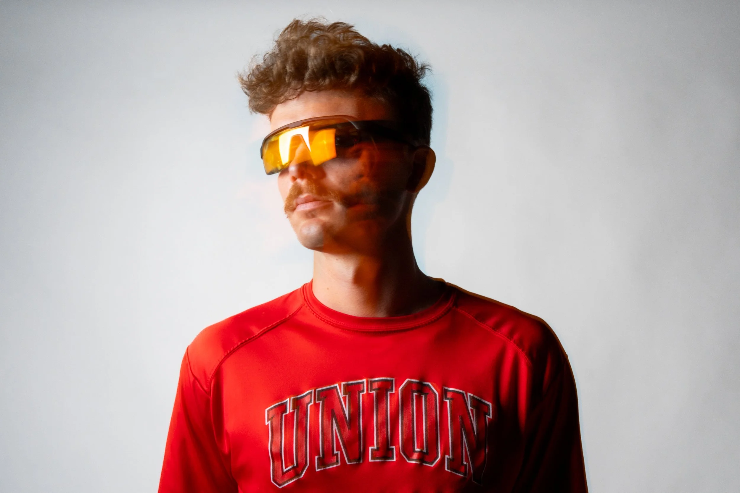 A young man with curly hair and a mustache wearing futuristic sunglasses and a red T-shirt