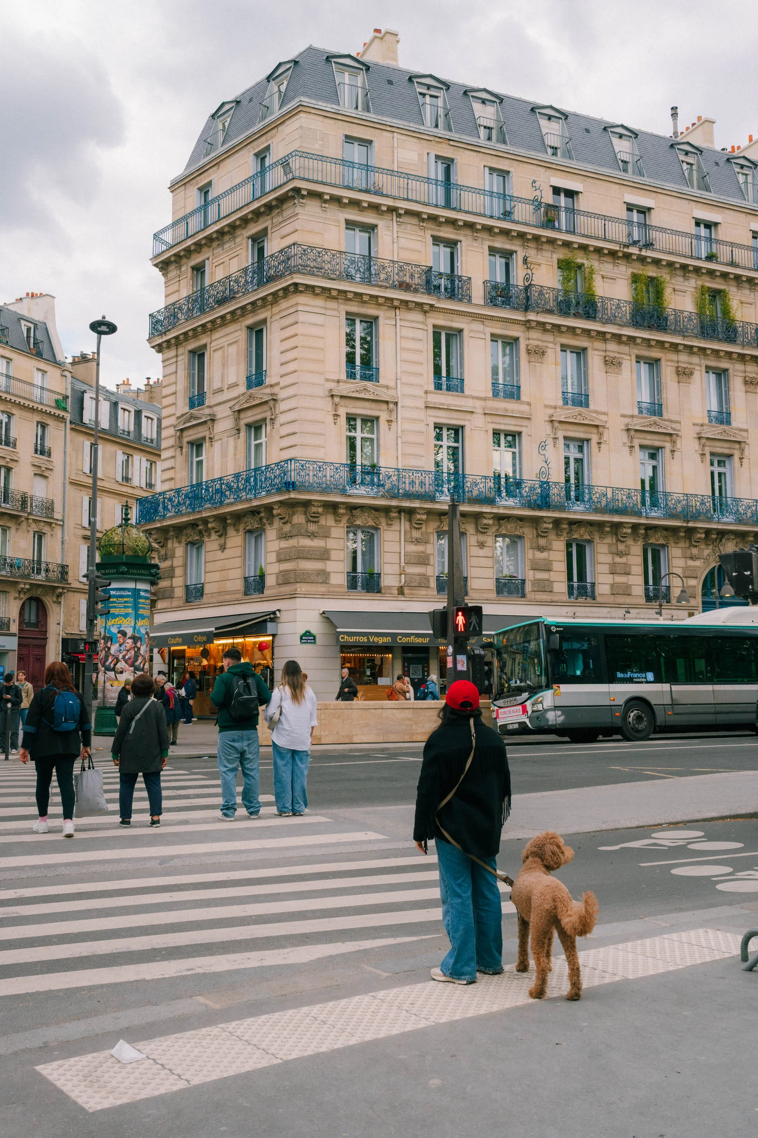 People waiting to cross the street at a busy intersection in Paris, France, in front of a classic Parisian building with wrought-iron balconies, a bus, street signs, and a cloudy sky.