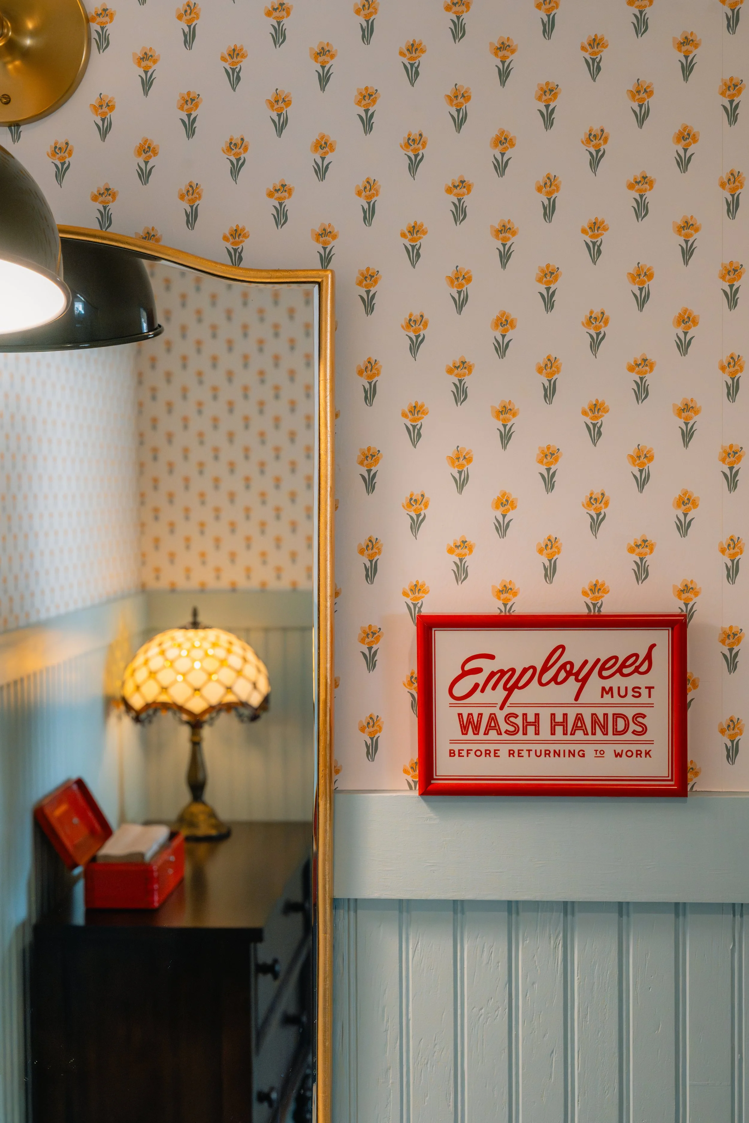 Interior of a vintage-style room with floral wallpaper, a mirror, a small black table with a lamp, and a red sign that says, "Employees must wash hands before returning to work."