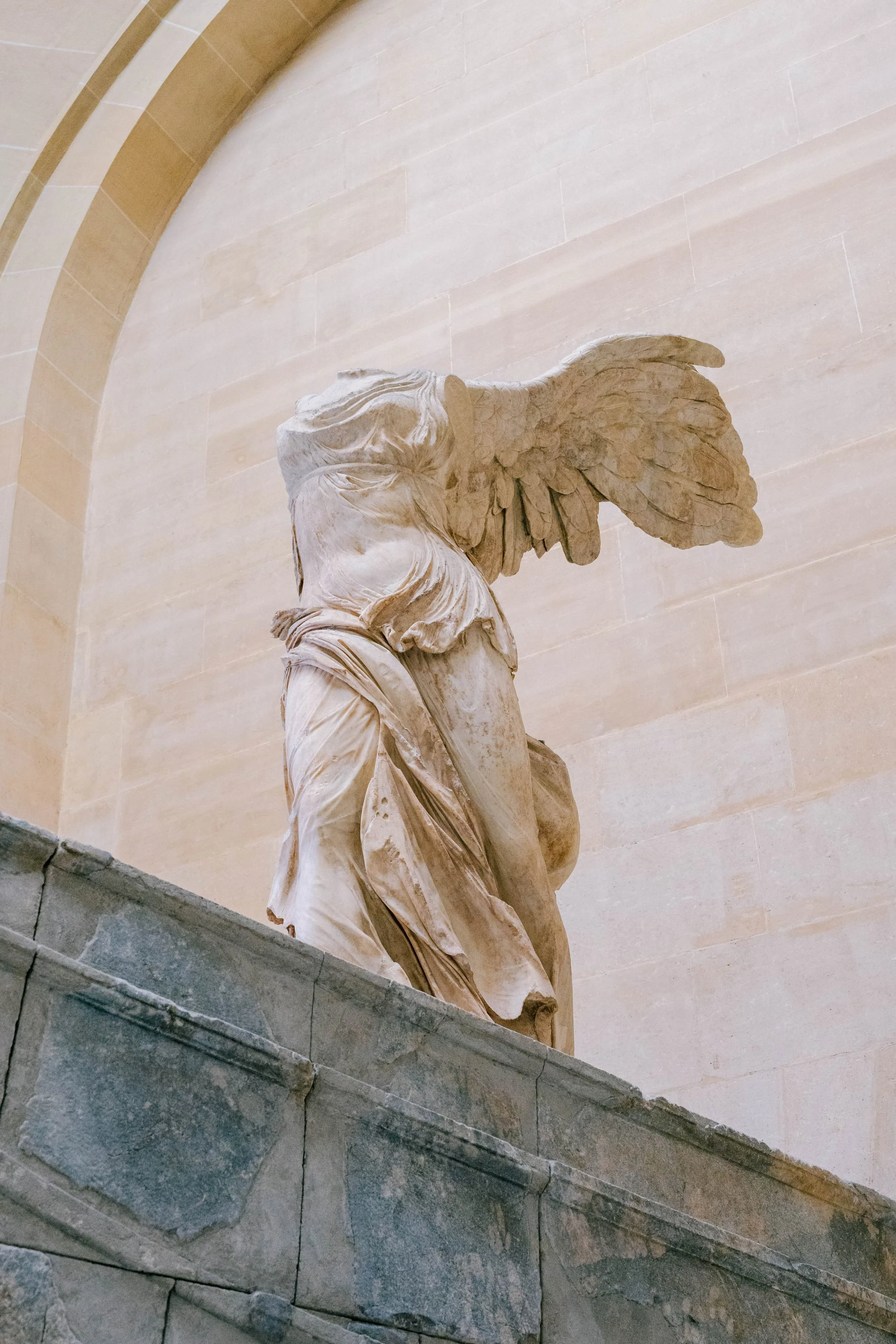 Close-up of the head and upper torso of a marble statue of Nike, the Greek goddess of victory, with wings, placed on a stone ledge against a beige wall with an arched architectural detail.