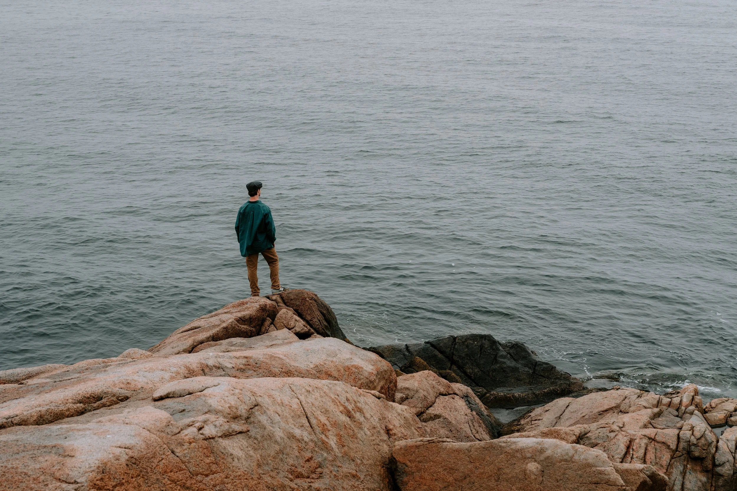 A man dressed in a dark green jacket, brown pants, and a cap standing on a rocky shoreline, looking out over the ocean
